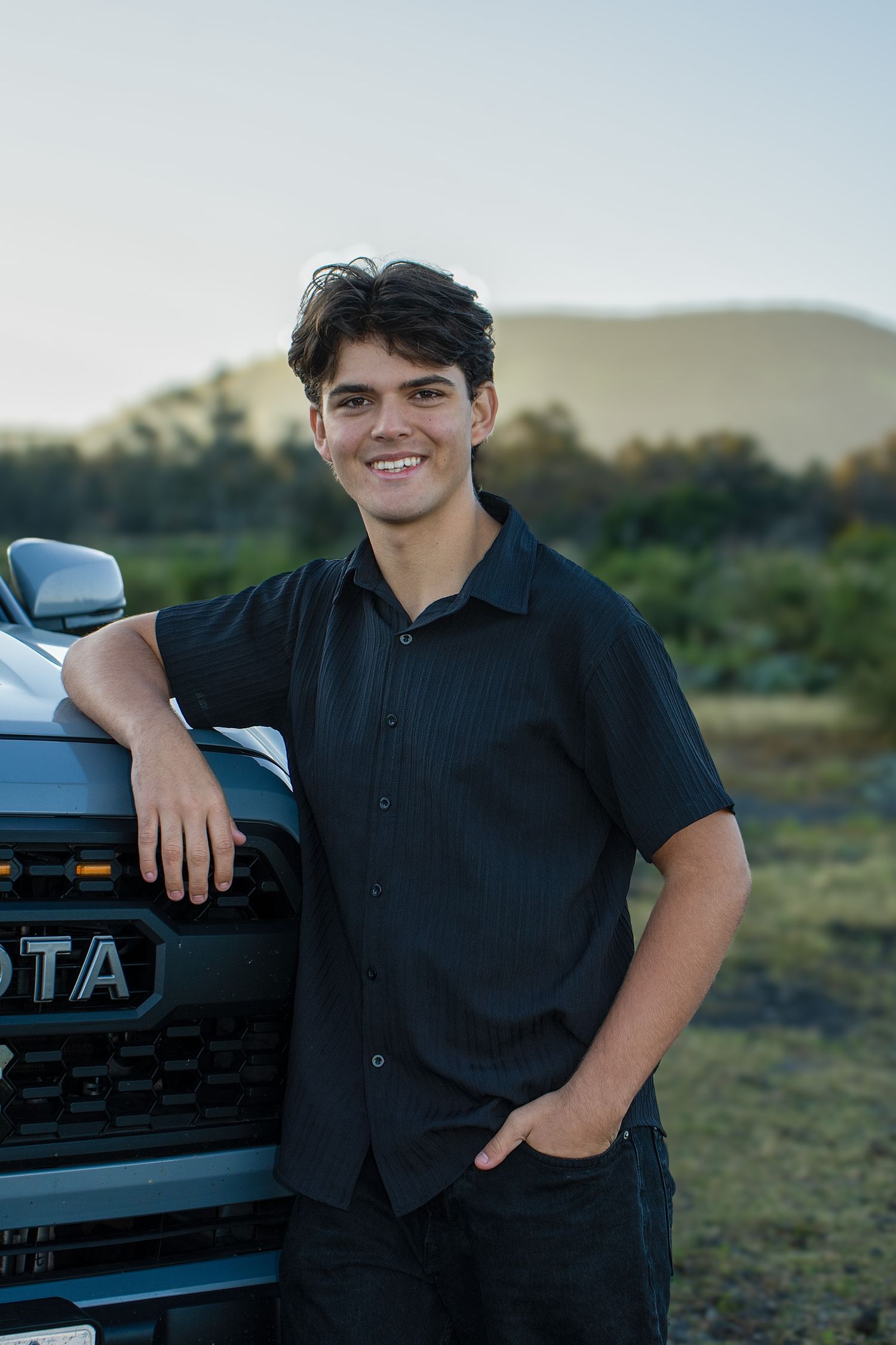 Young man in black shirt smiling beside a Toyota truck with mountain scenery in the background.