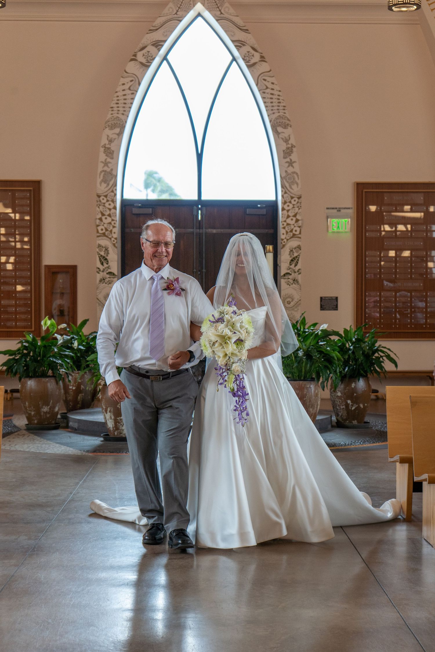 Bride in white gown and veil walks down church aisle with escort holding bouquet of white and purple flowers.