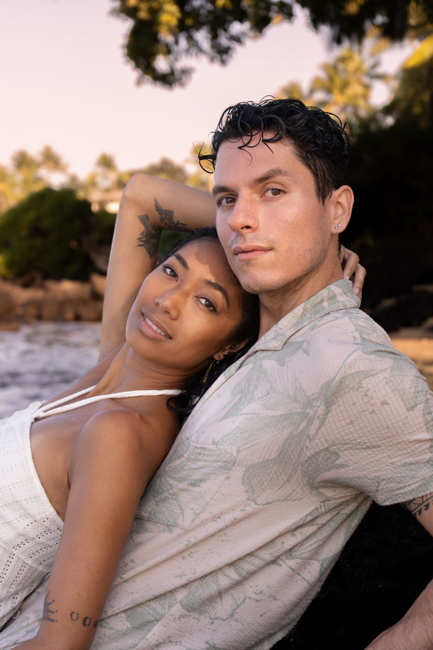 A couple sits together at sunset on a tropical beach, the woman leaning against the man in a tender embrace.