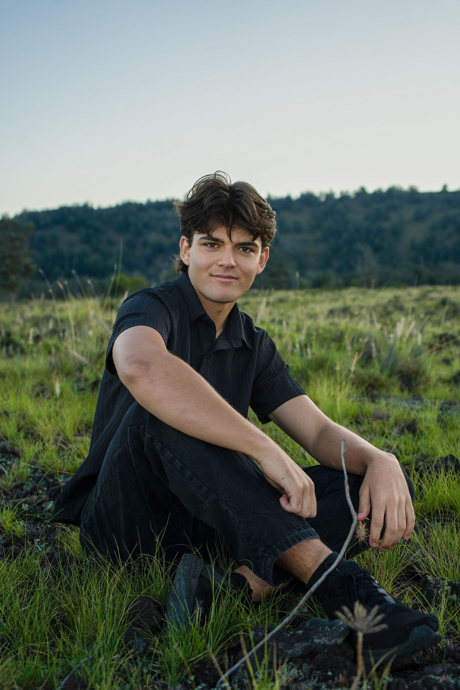 A person wearing black sits casually in a grassy field with trees visible in the background during golden hour.