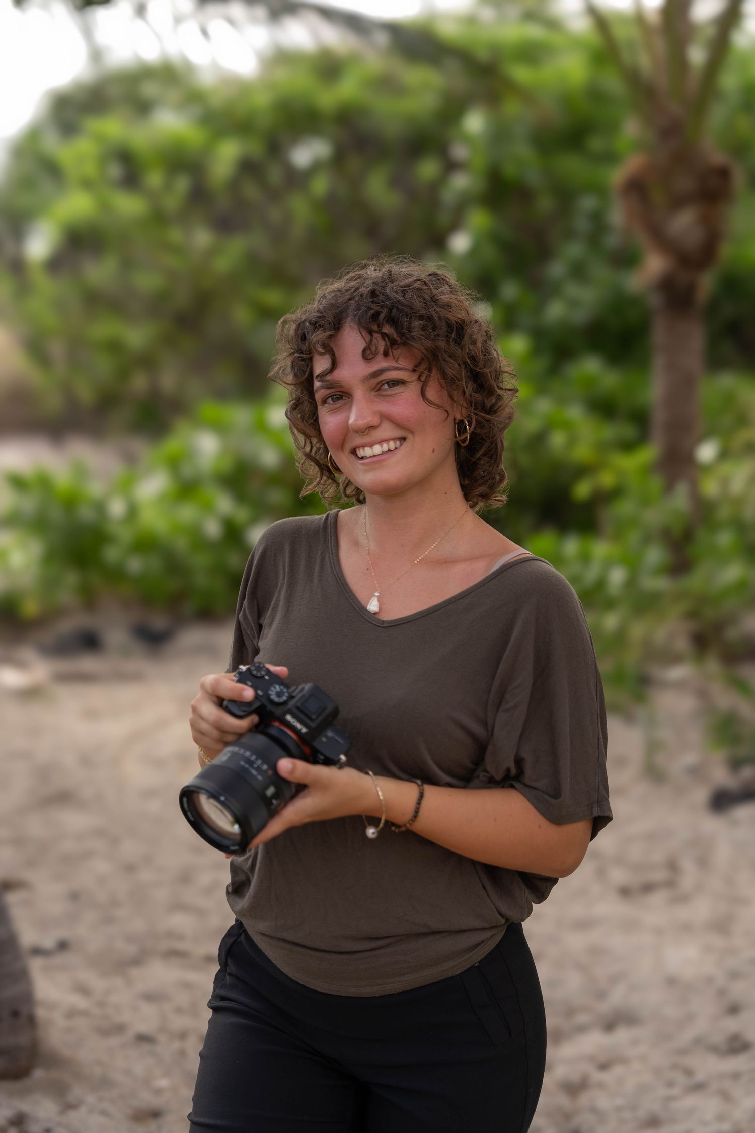 Smiling woman with curly hair holding a DSLR camera outdoors, wearing an olive green top with green trees behind her.