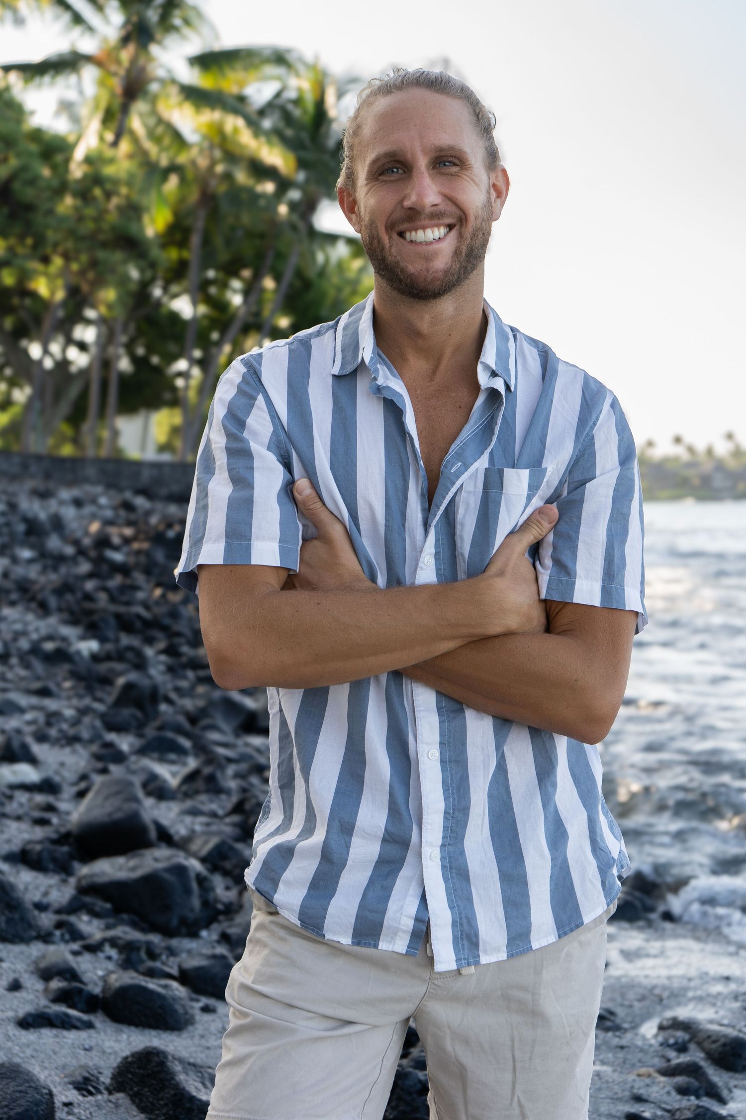 Man in striped shirt smiling on black rock beach with palm trees.