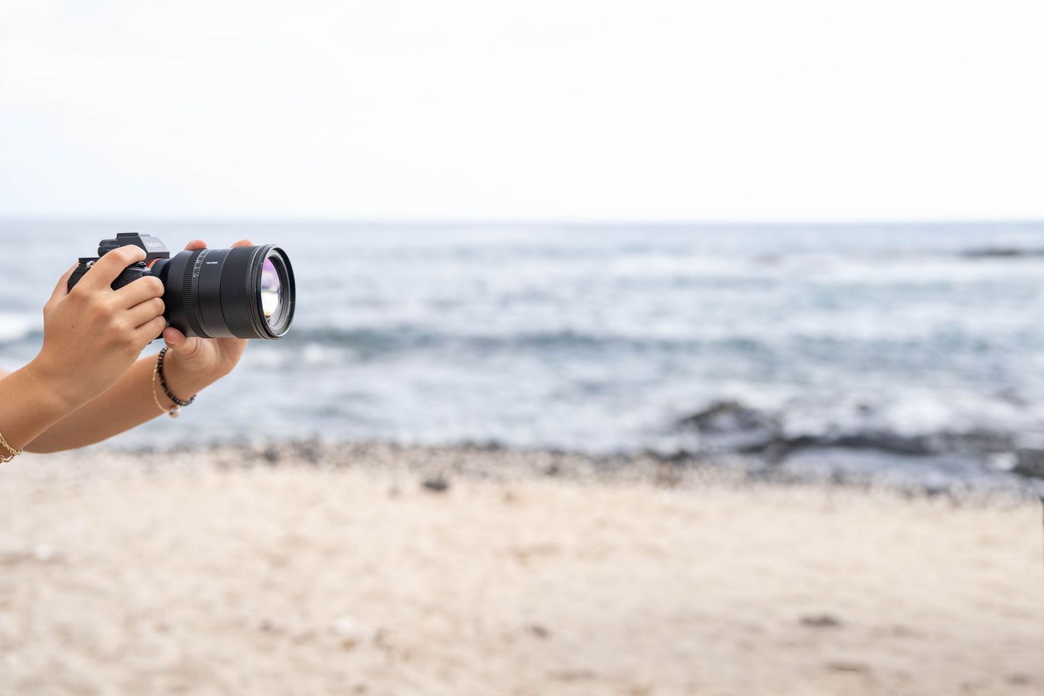 Hand holding a DSLR camera on a sandy beach with ocean waves in the background on a cloudy day.