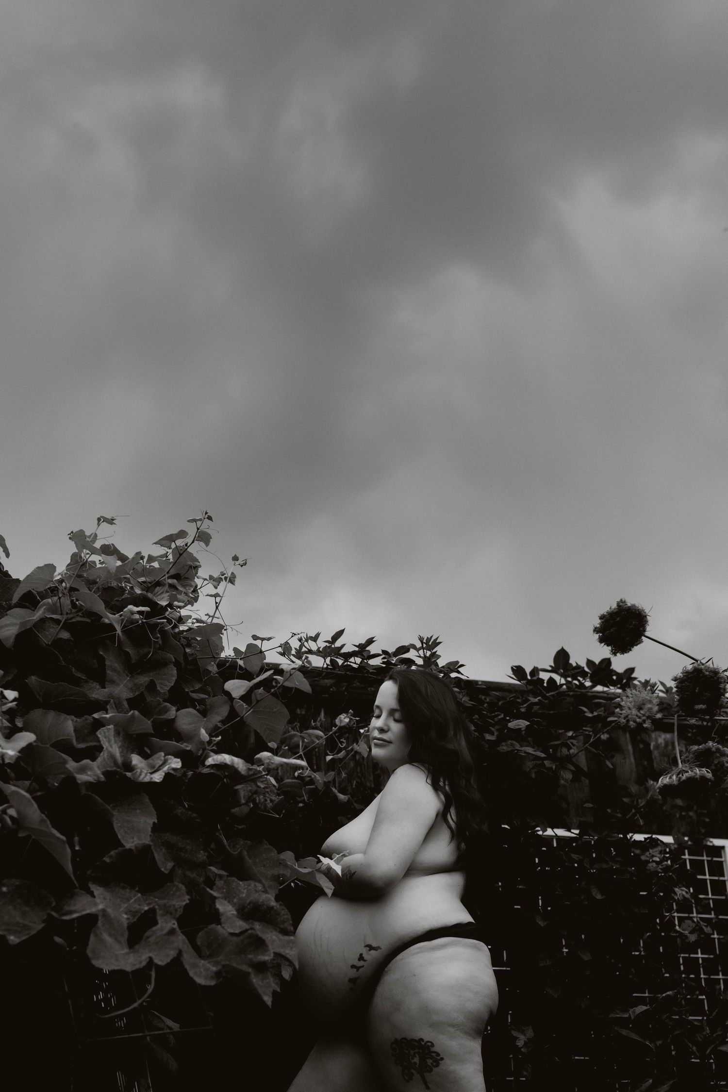 Black and white outdoor maternity portrait of a pregnant woman standing behind foliage under a dramatic cloudy sky.