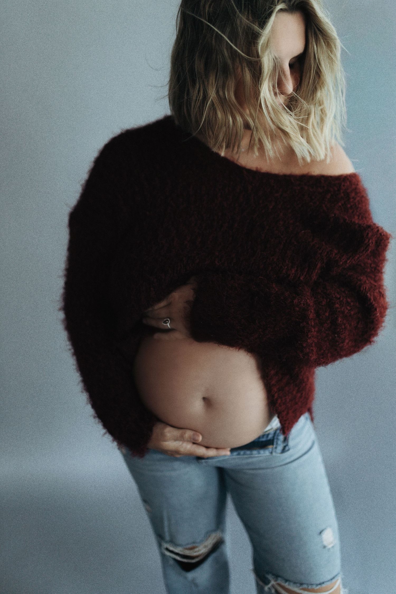 A person poses in a cropped burgundy sweater and distressed light blue jeans against a gray background.