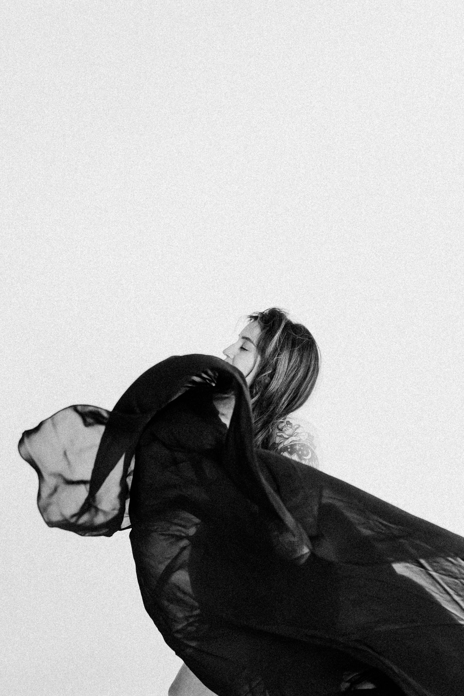 A black and white artistic photograph shows a figure in flowing dark fabric moving dramatically against a light background.