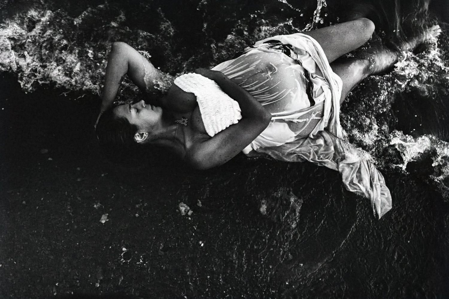 Black and white artistic photograph of a figure reclining on dark textured ground.
