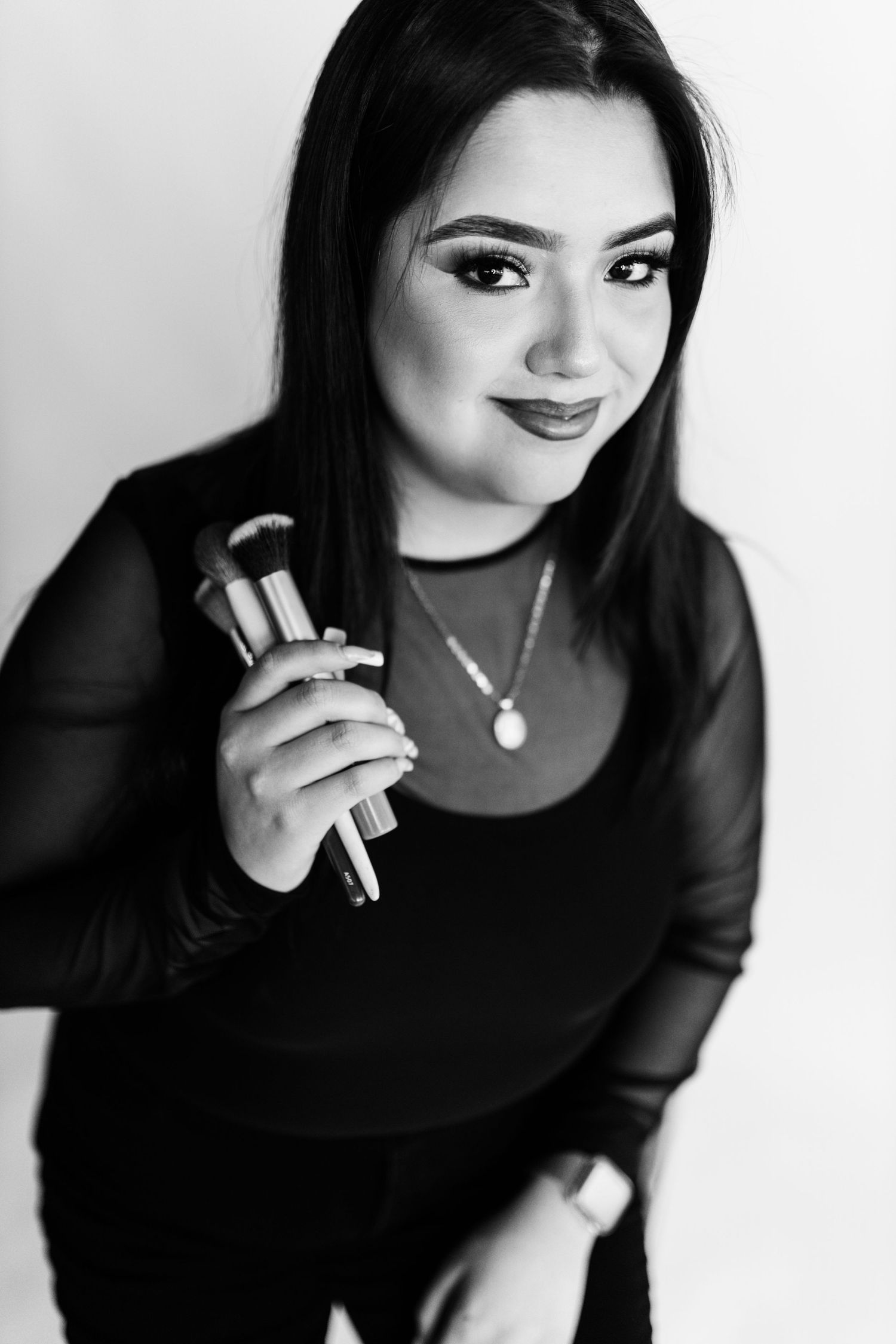 Black and white portrait of Elysse Jimenez, Dallas makeup artist for White Moth Gallery, holding makeup brushes.