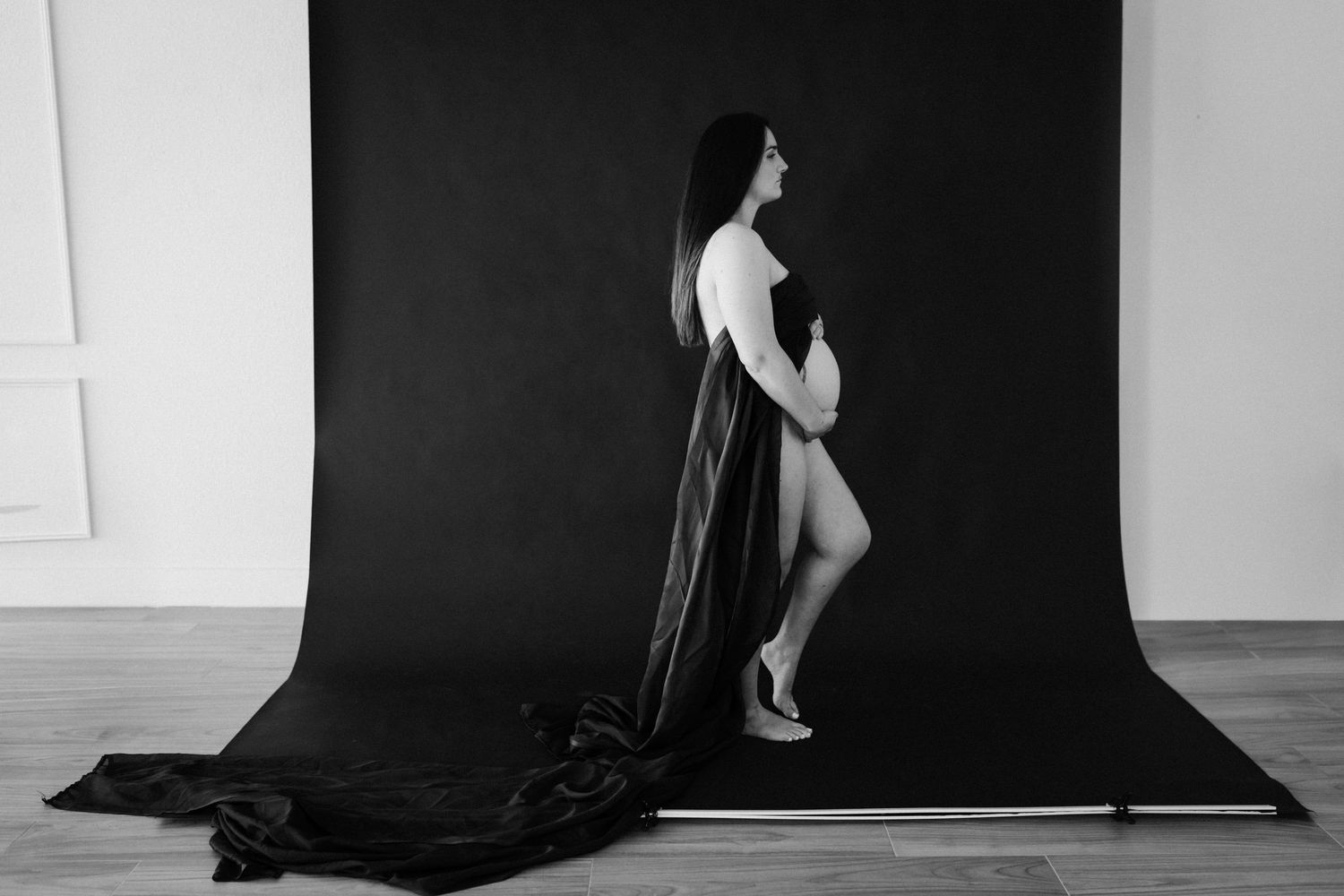 Black and white maternity photo of a pregnant woman posing gracefully with a flowing dark fabric on a studio backdrop.