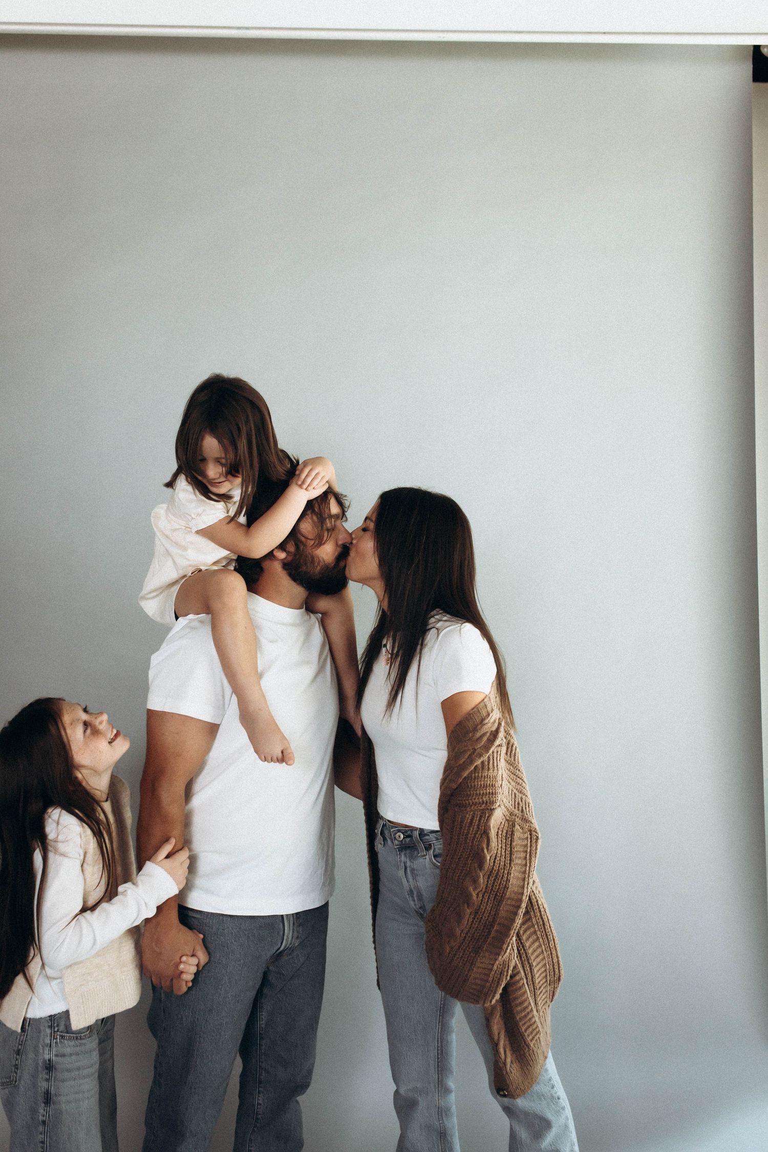 A family wearing white tops and gray pants poses together against a light gray wall.