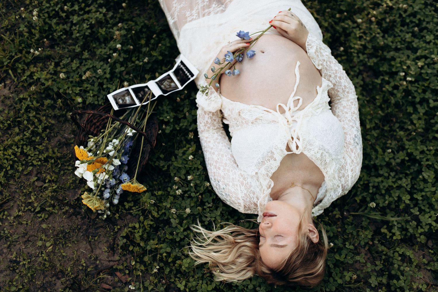 Pregnant woman in white lace dress lying on grass with wildflowers and ultrasound photos beside her.