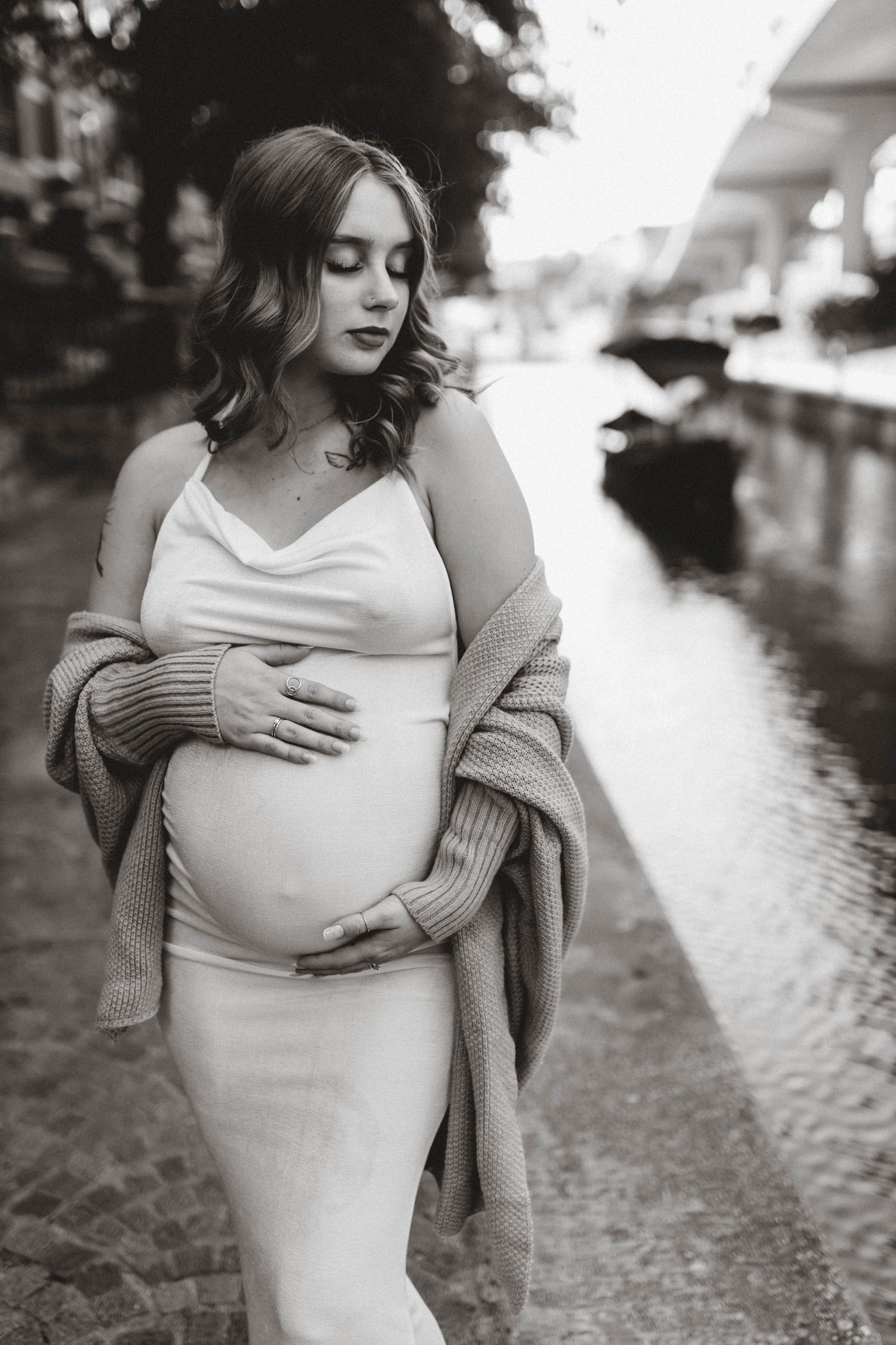 A black and white maternity portrait of an expectant mother in a white dress by a canal.