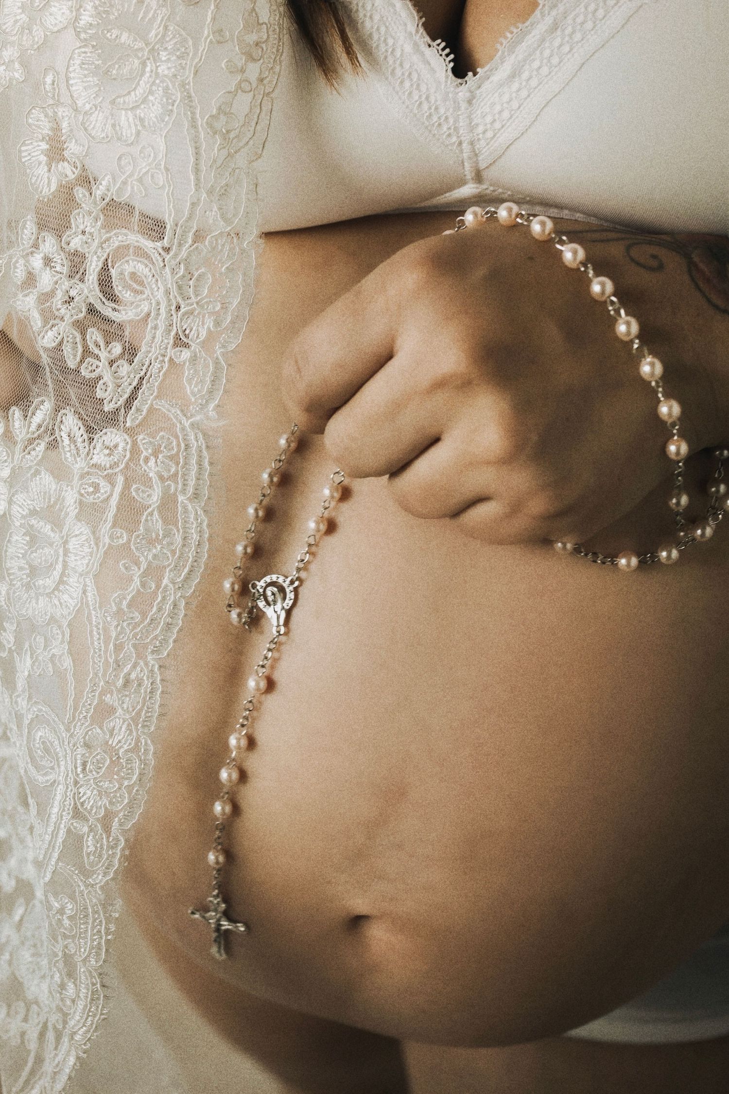 A delicate pearl rosary rests on a pregnant belly next to white lace wedding dress details.
