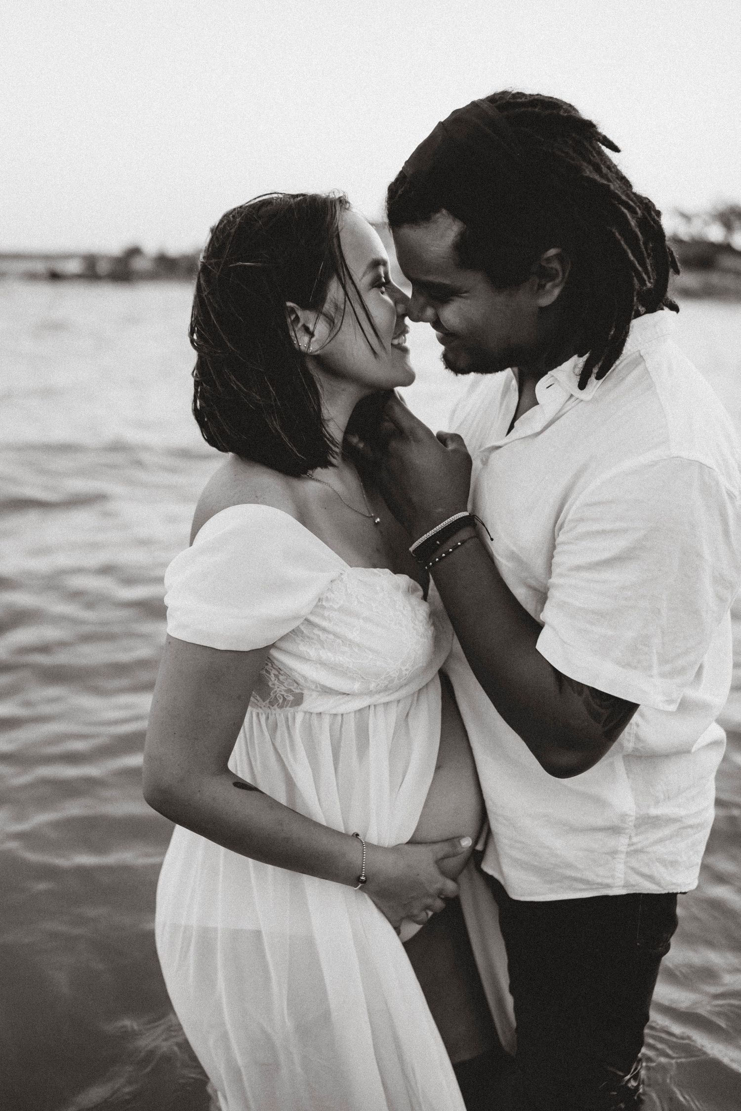An intimate black and white portrait of an expecting couple sharing a tender moment by the water.