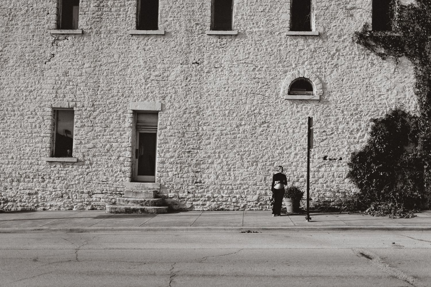 A solitary figure sits on a bench against a stark white stone building with rectangular windows.