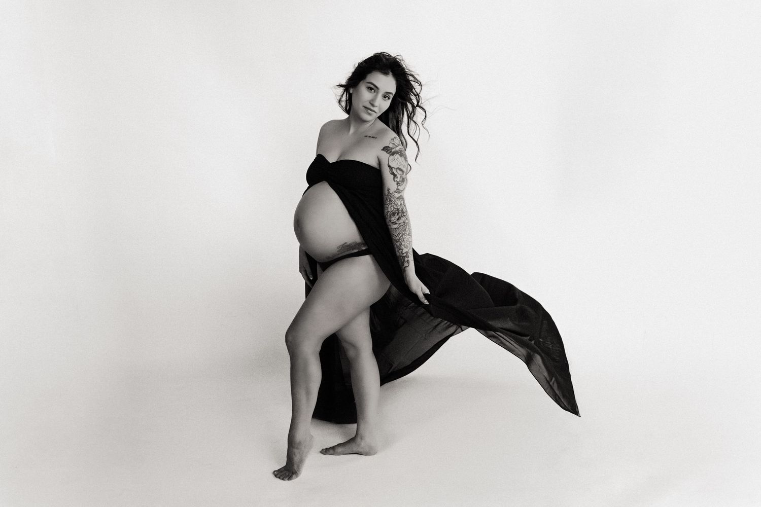 Artistic black and white maternity photo showing an expectant mother in flowing fabric against white background.