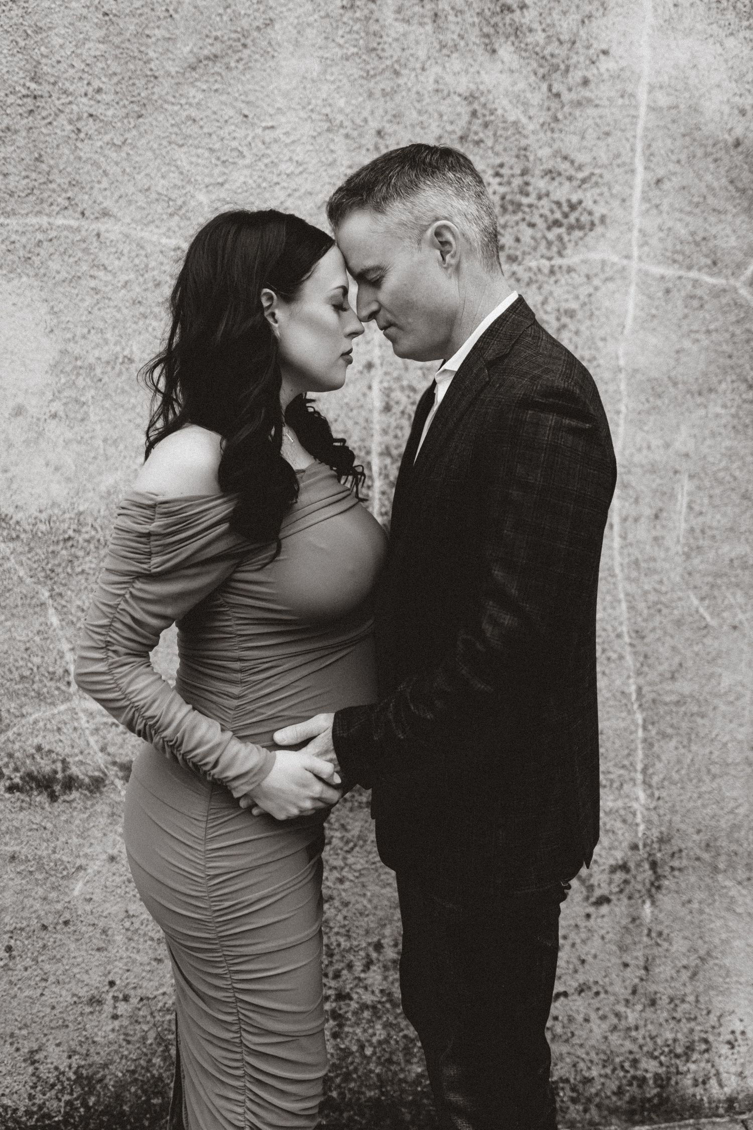A romantic maternity photoshoot shows an intimate couple embracing against a textured wall background.