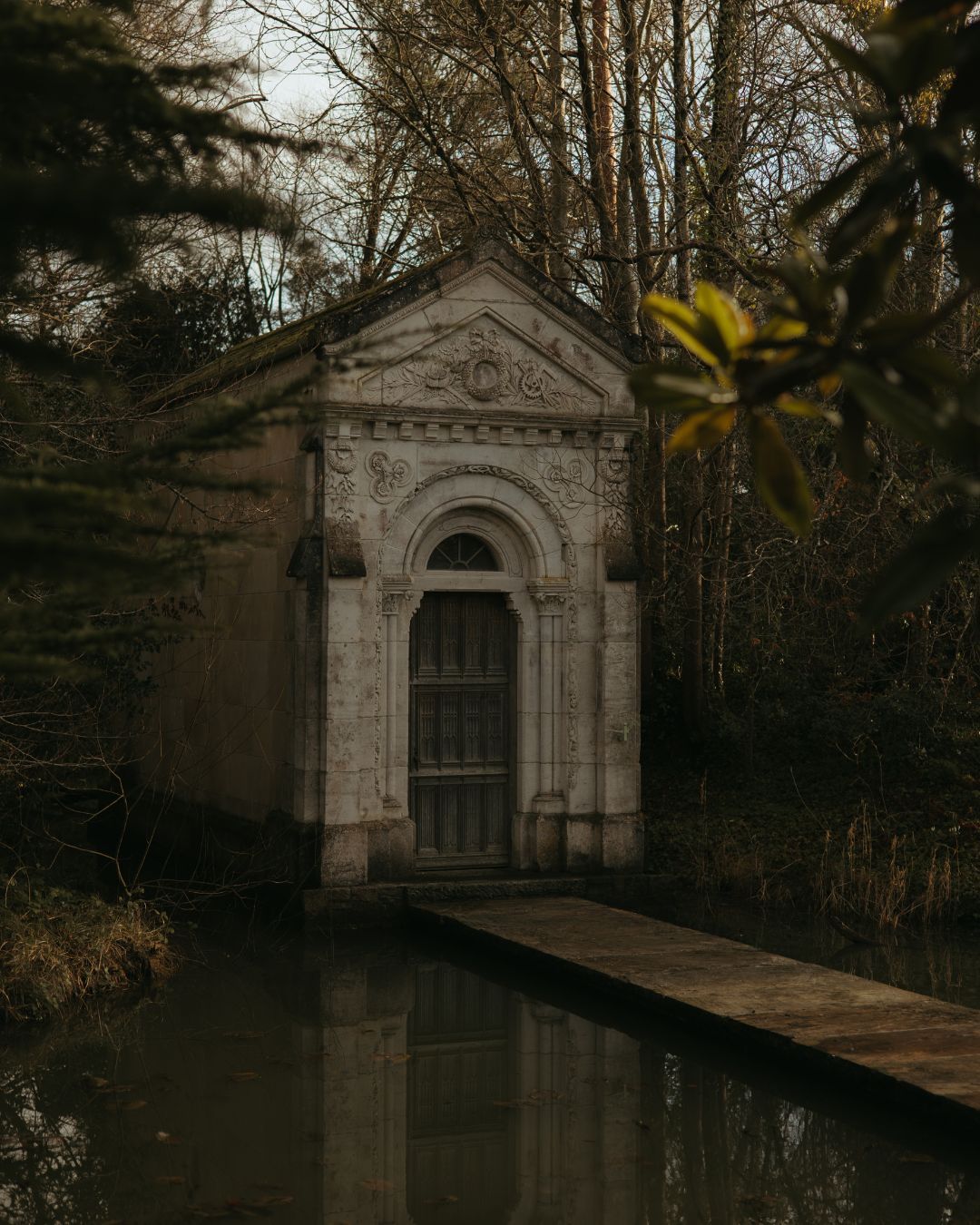 A Gothic stone mausoleum with ornate arched entrance reflects in still water surrounded by bare winter trees.