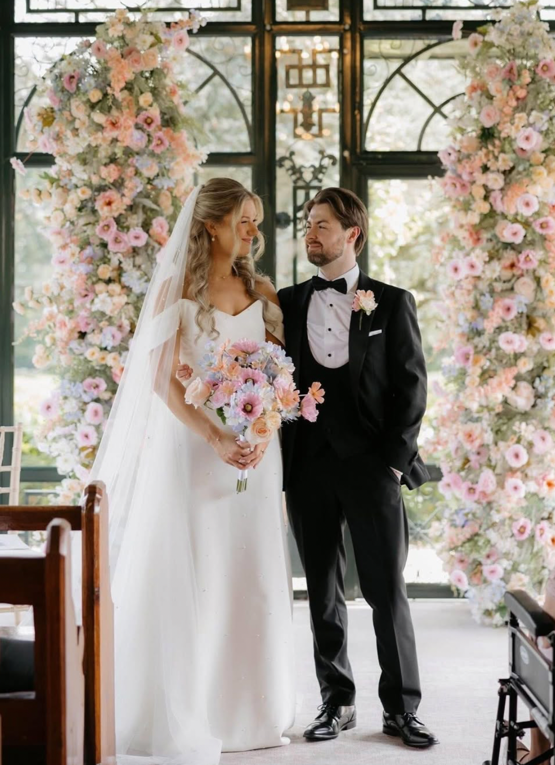 A couple shares a tender moment in a wedding venue decorated with pink floral arches and natural lighting.