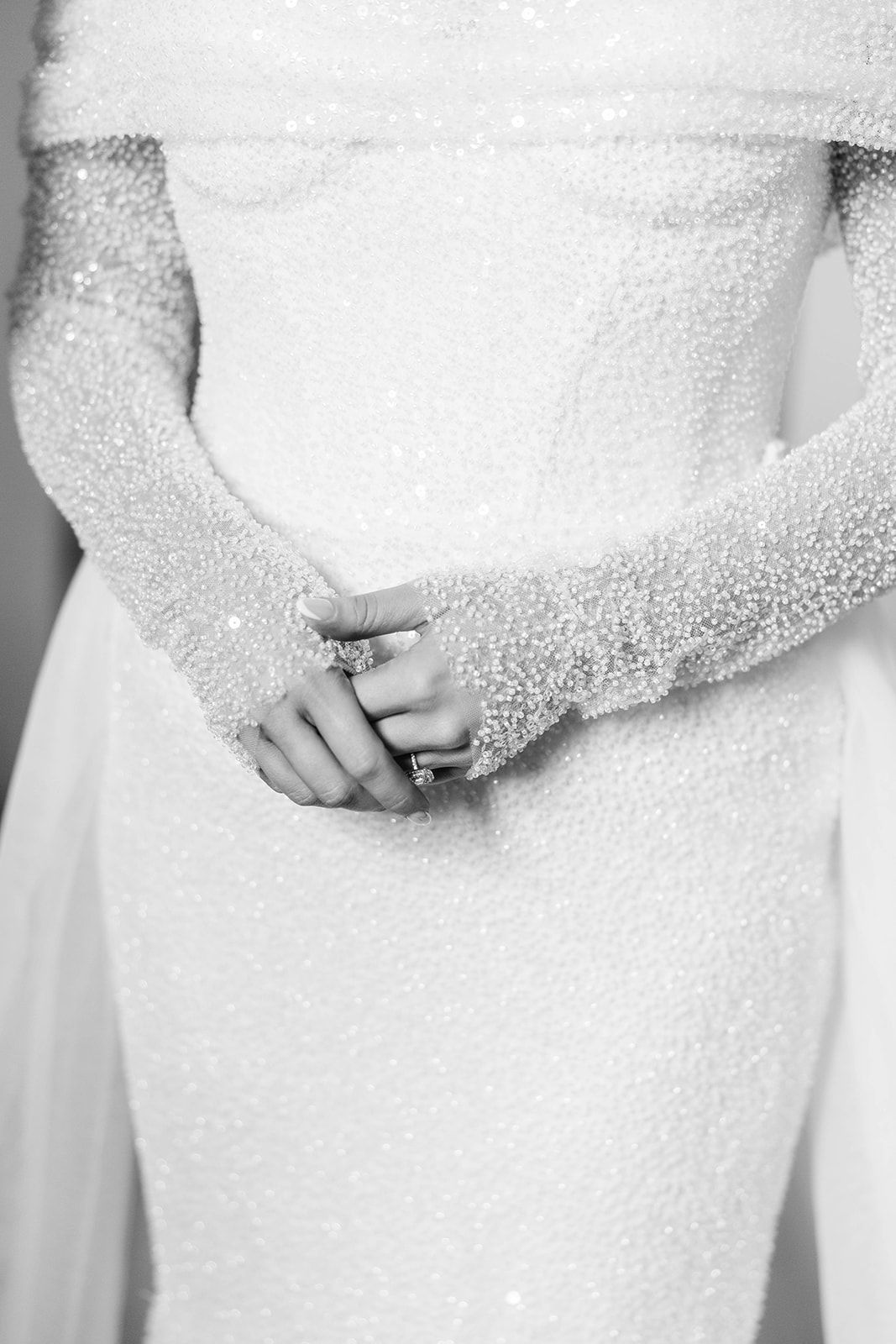 Close-up black and white photo of hands clasped together wearing long lace sleeves and a fitted white wedding dress.
