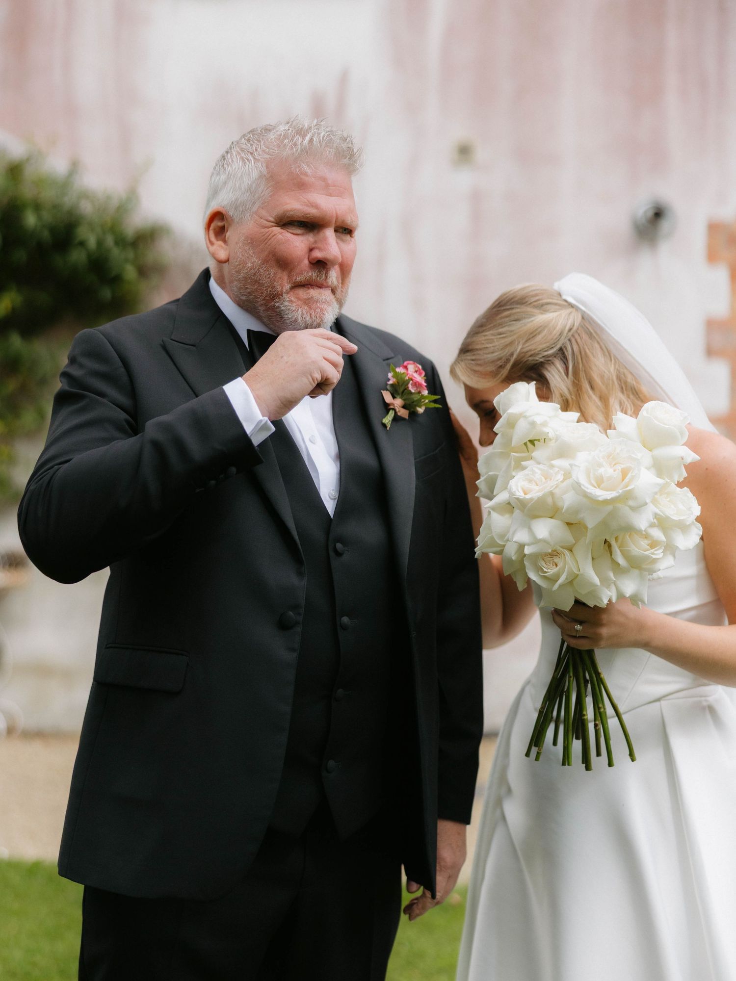 Wedding couple shares an emotional moment with white bouquet against outdoor backdrop.