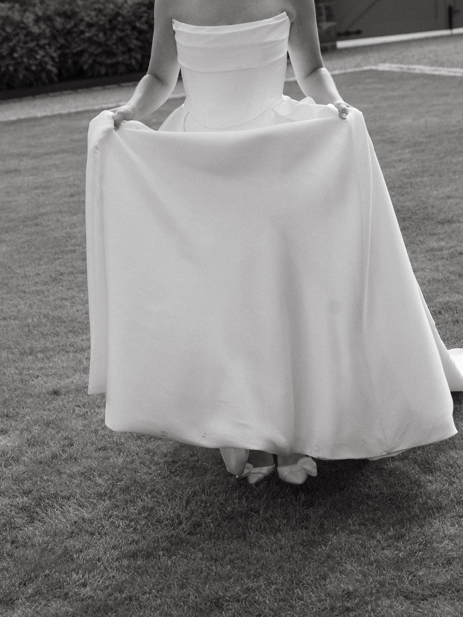 Black and white photo showing the flowing hemline of a wedding dress on grass.