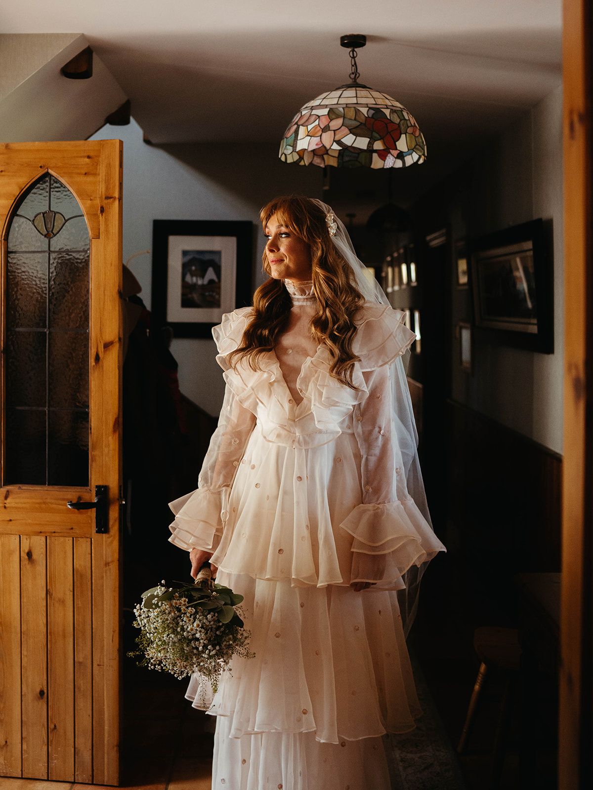 A bride in a romantic tiered white wedding dress stands holding a bouquet under a vintage pendant light.