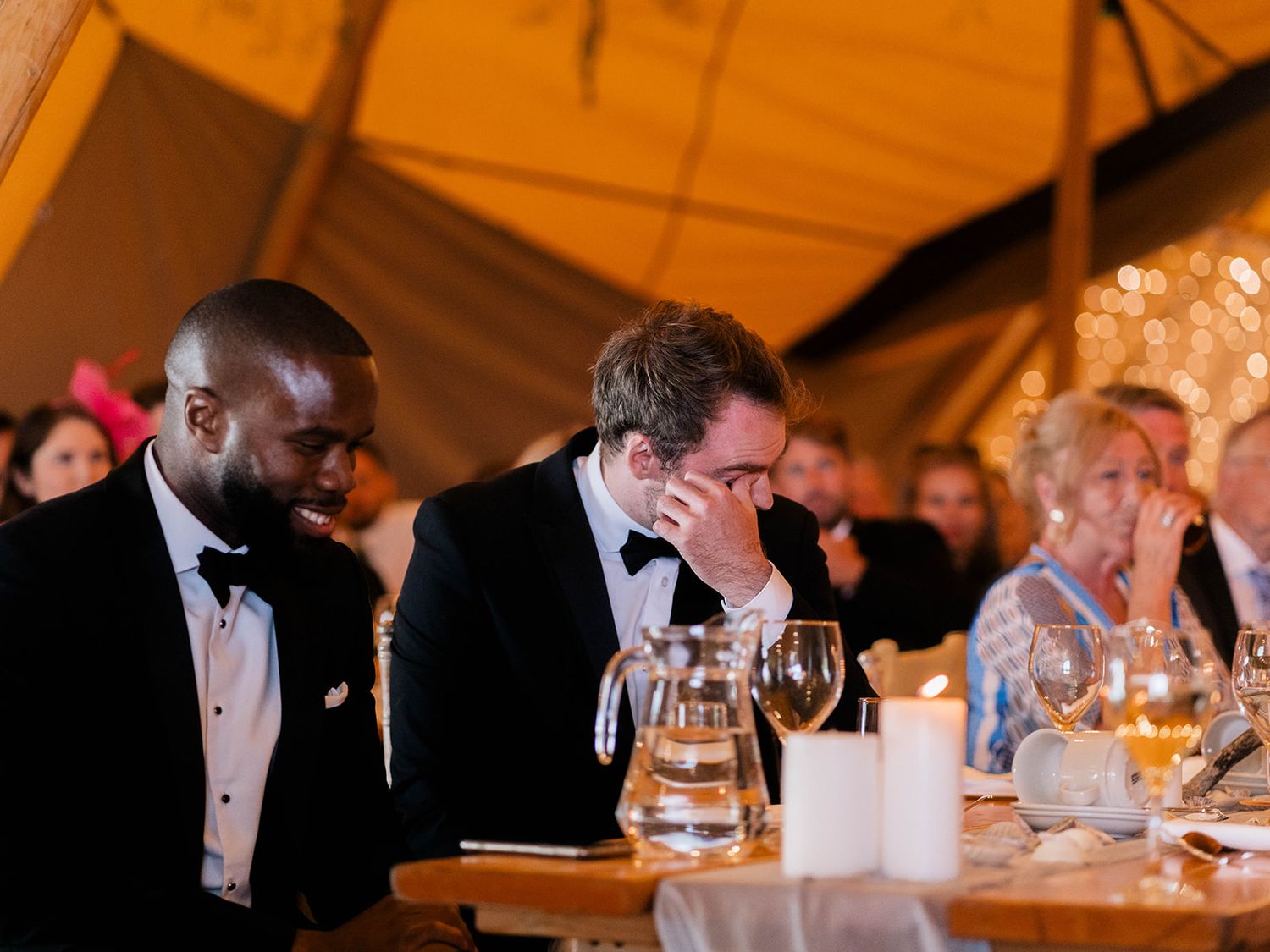 Wedding guests showing emotional reactions during speeches at a reception dinner under tent lighting.