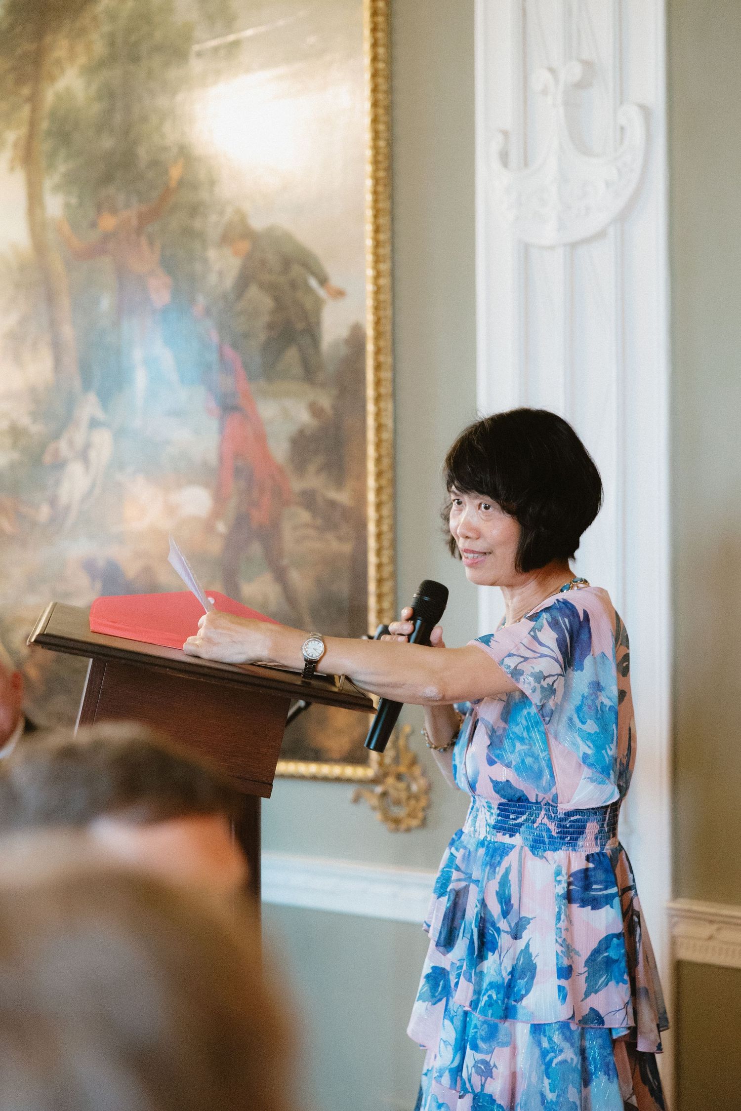 Speaker at podium delivers speech in elegant room with classical painting and white moldings.
