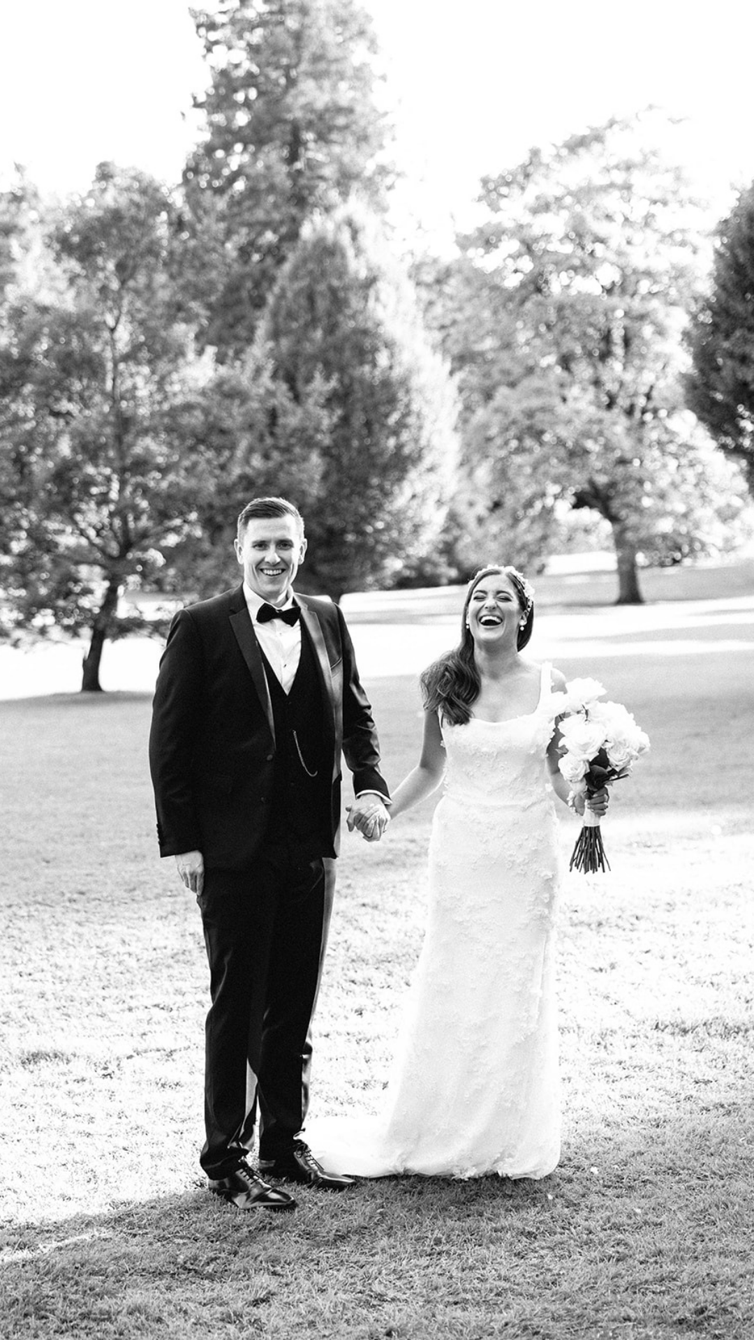 Black and white wedding photo of couple laughing together on manicured lawn.