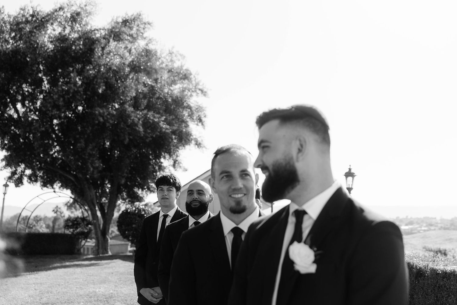 Black and white photo of suited guests at an outdoor wedding ceremony with trees and nature in the background.