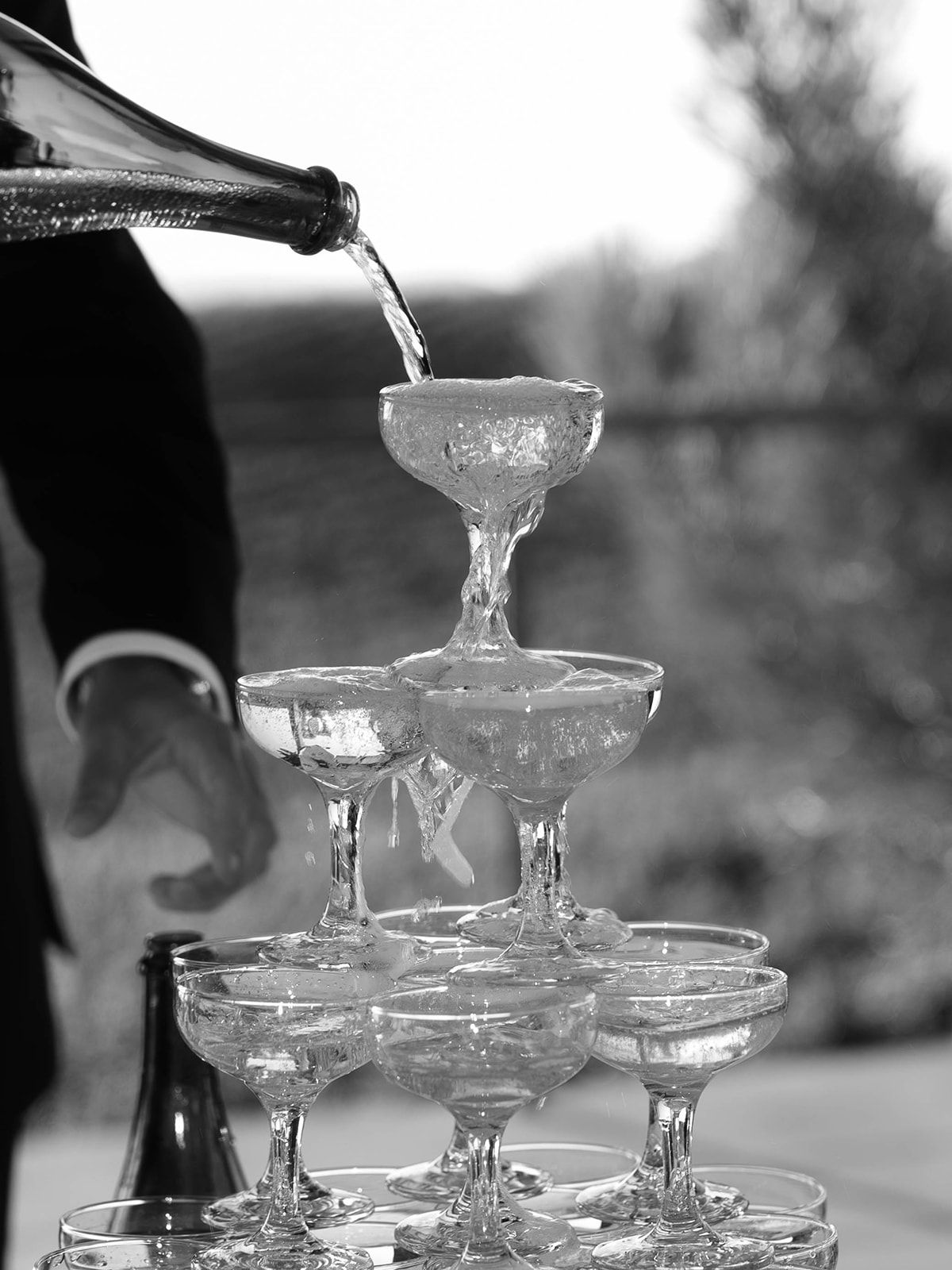 Champagne being poured into a tower of stacked coupe glasses in black and white photography.
