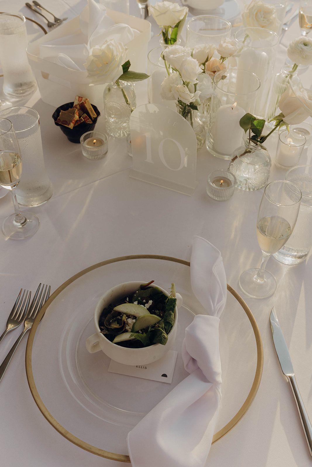 Elegant white and gold wedding table setting with floral centerpieces, wine glasses, and a plated salad course.