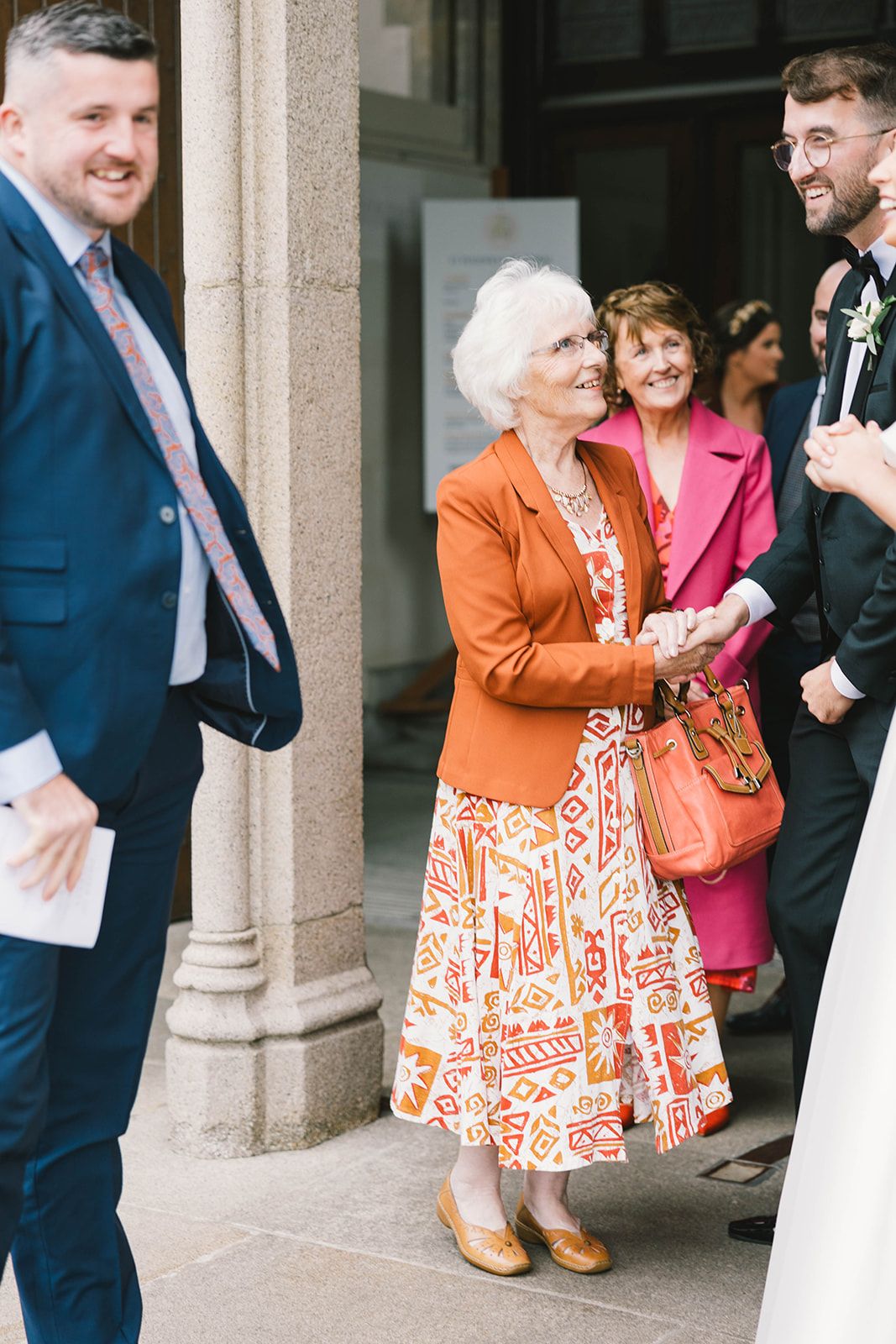 A group of people conversing outside a building, with someone wearing a bright orange and white patterned dress with matching jacket.