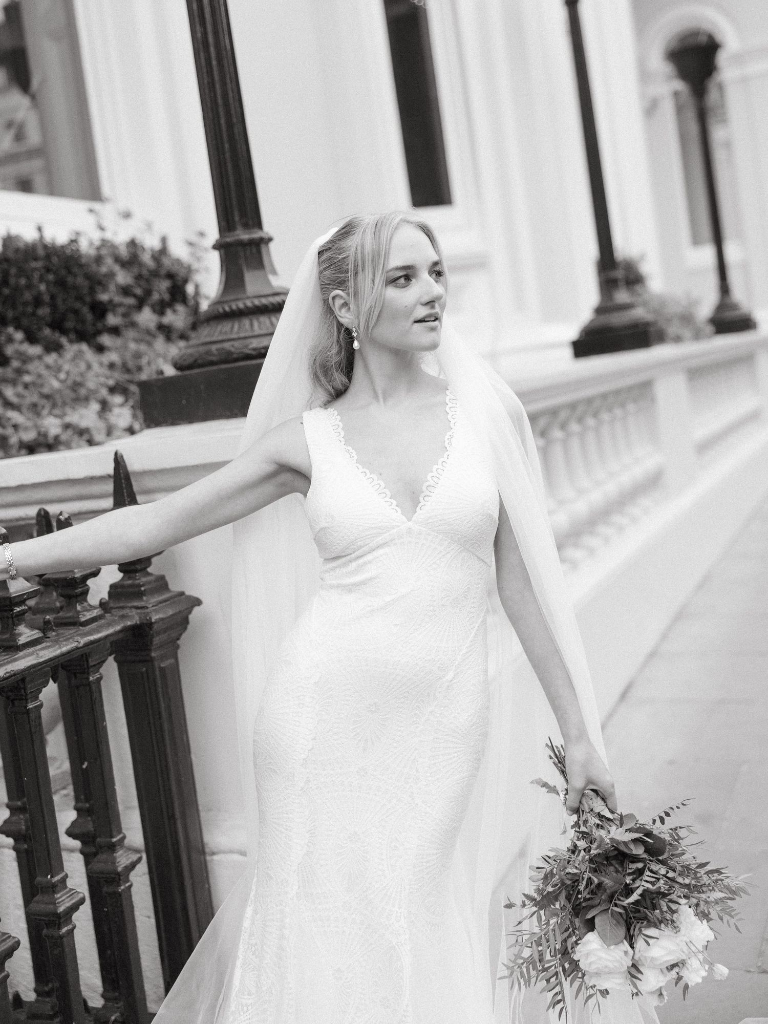 A bride in an elegant white dress with a billowing veil poses against a white building exterior.