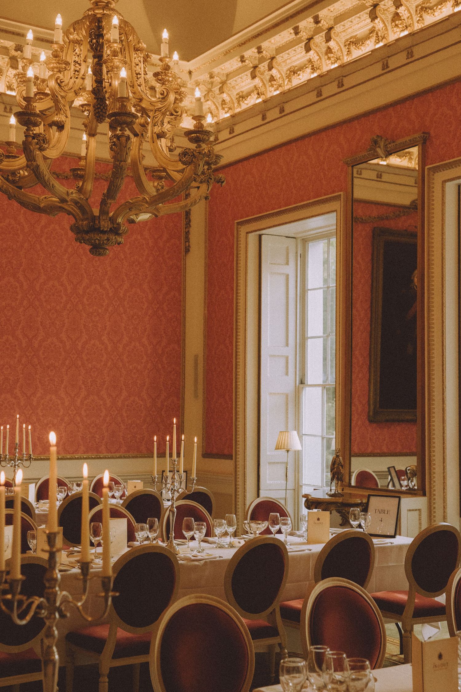 Elegant dining room with ornate chandeliers, red damask wallpaper, dark wooden chairs and classic table settings.