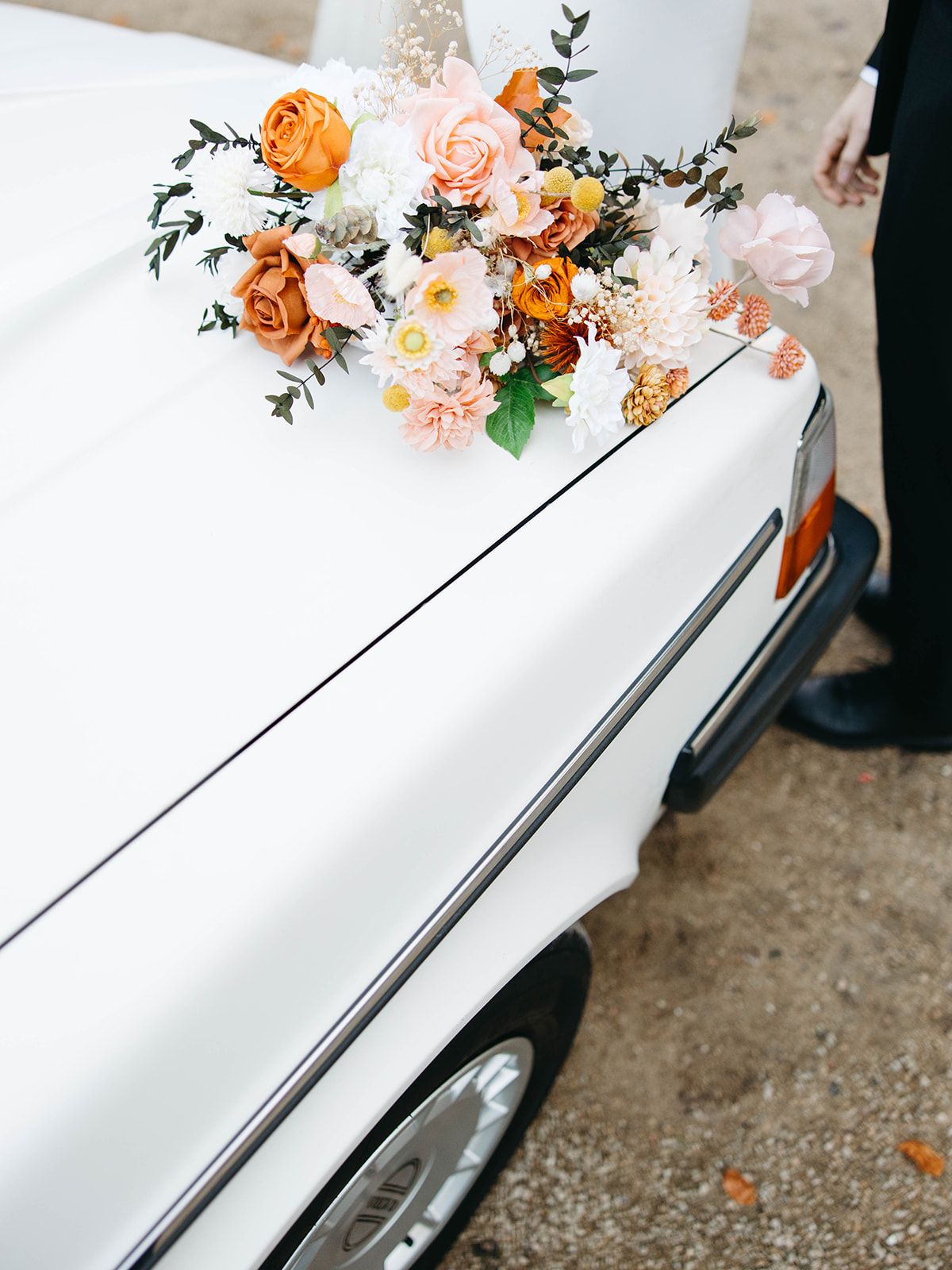 A vintage white car decorated with peach and orange floral arrangements on its hood, captured at a wedding event.