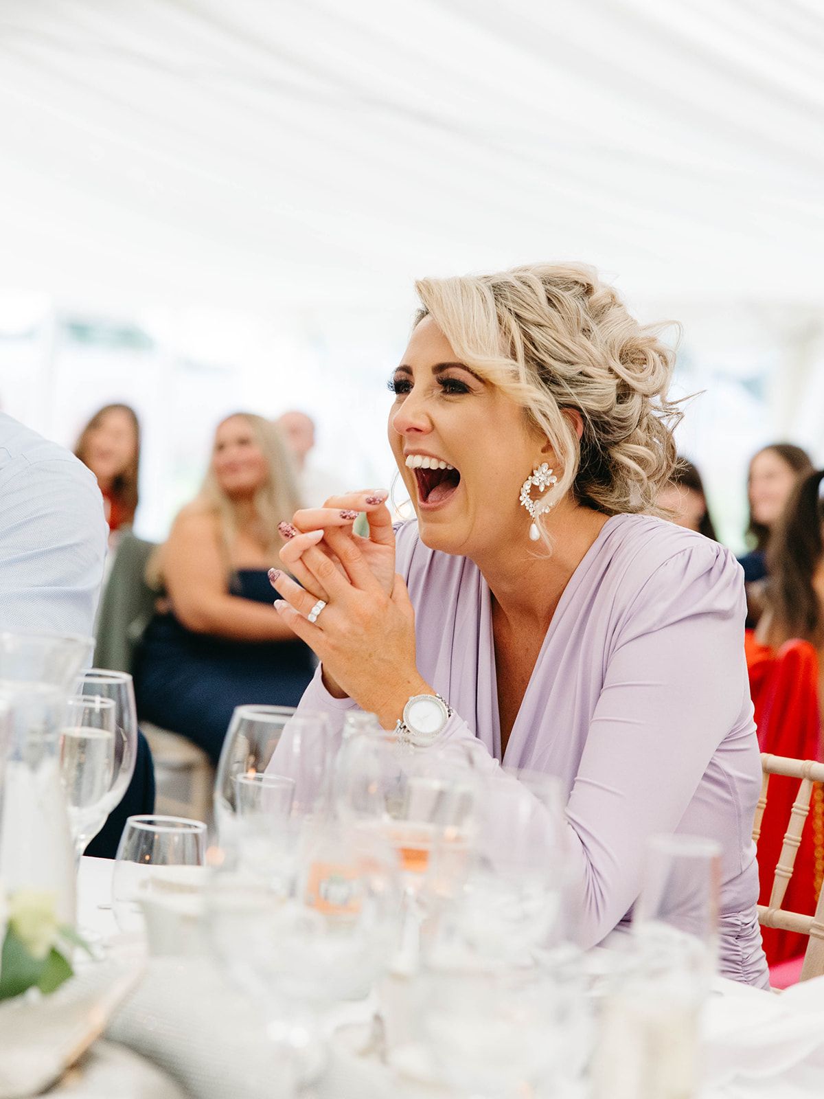 Event guest in lavender outfit laughing joyfully at dining table with wine glasses in elegant party setting.