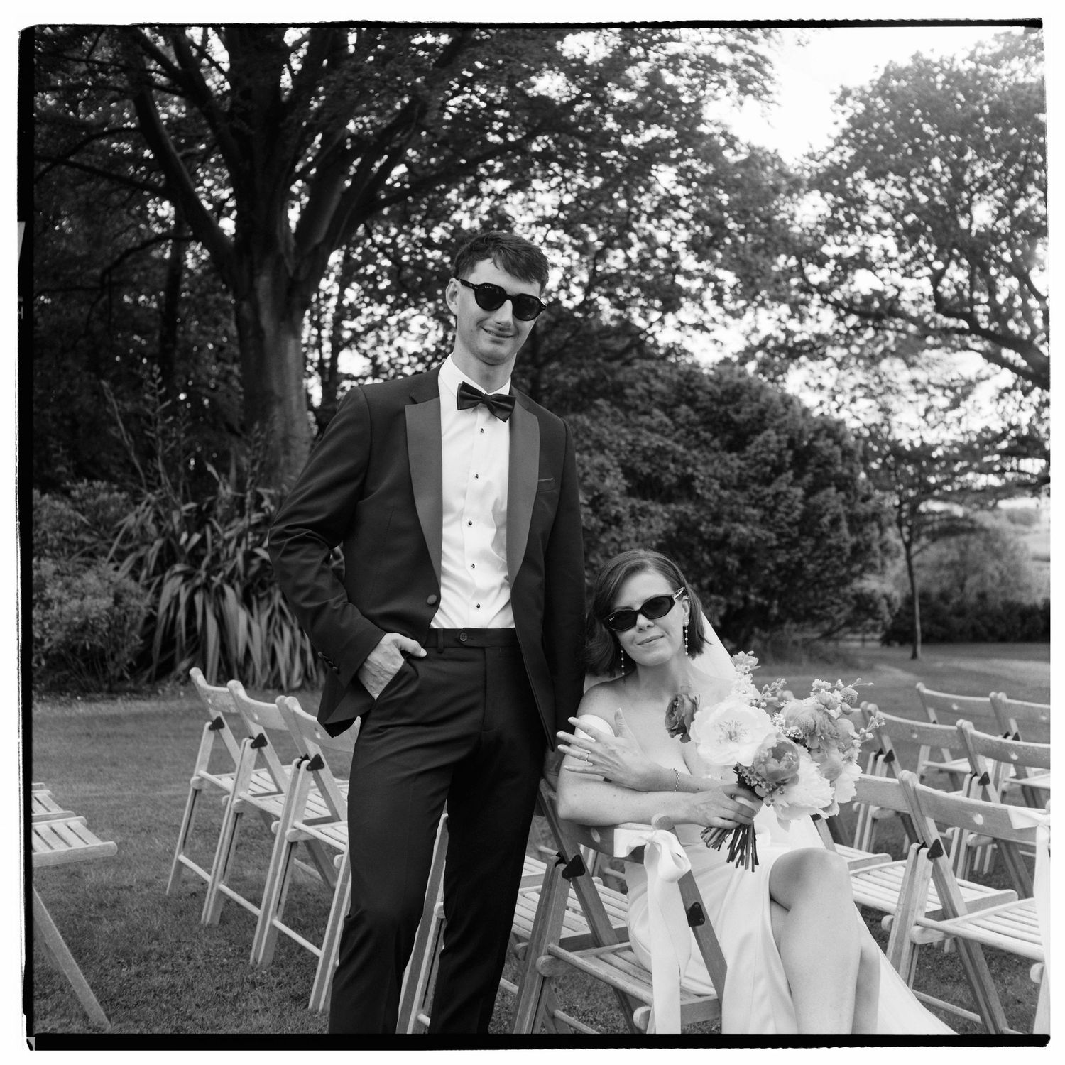 Black and white photo of a bride and groom in sunglasses posing outdoors at a garden wedding ceremony.