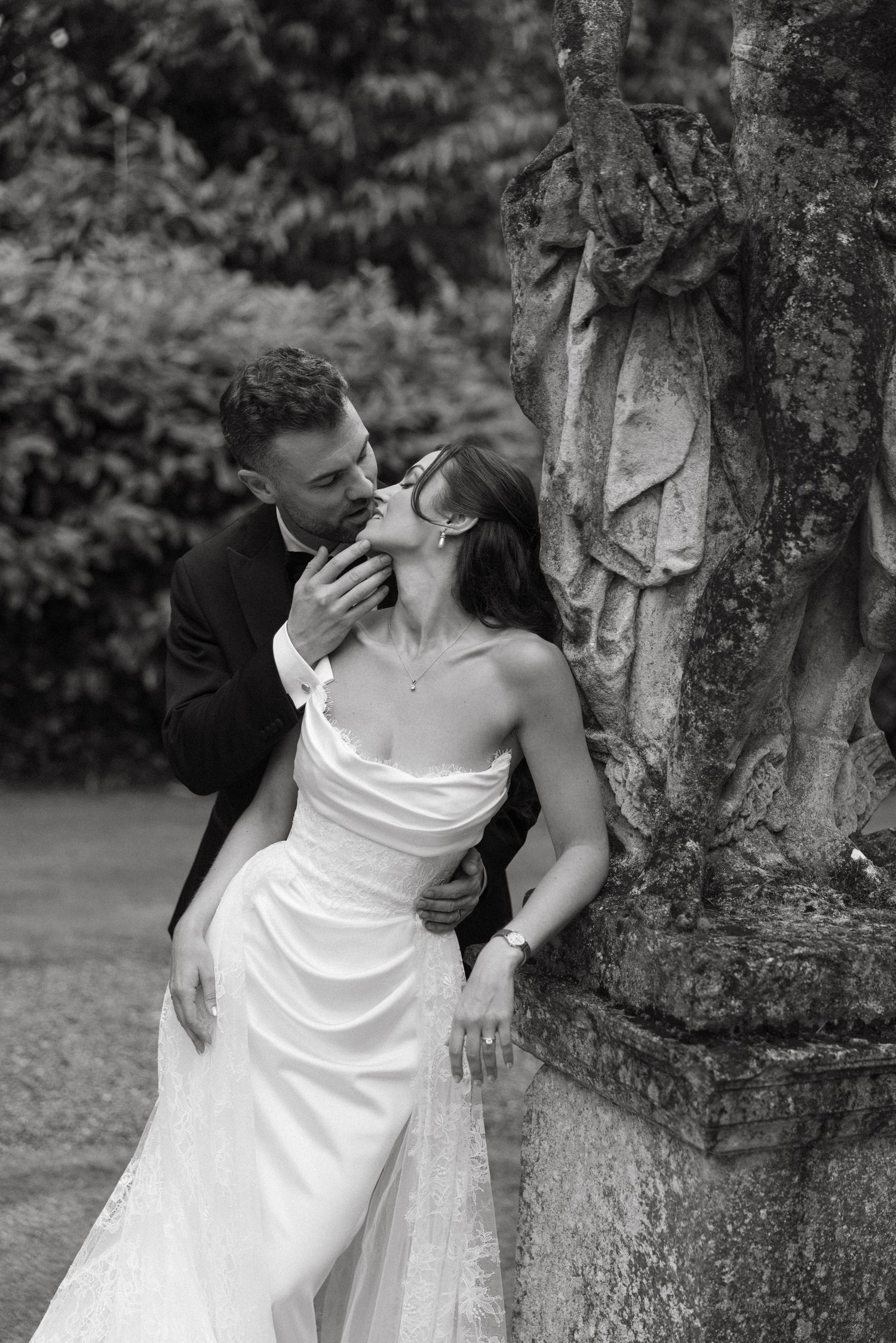 Black and white photo of a bride and groom sharing a romantic kiss beside an ornate stone sculpture outdoors.