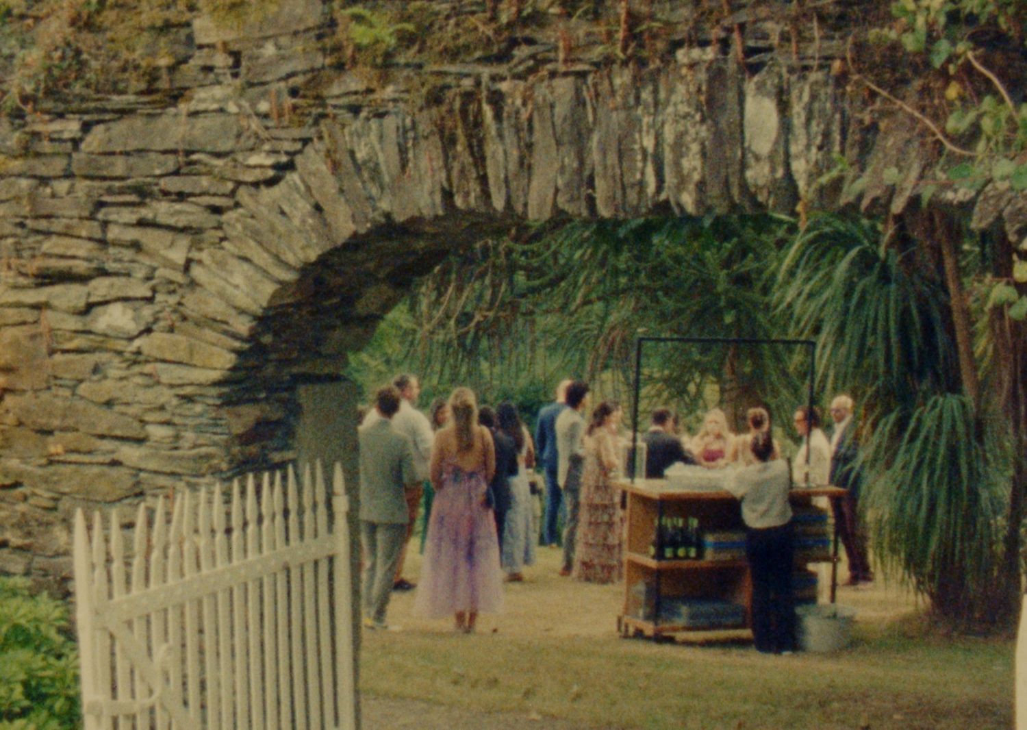 Guests gather under a stone archway at an outdoor garden party with lush greenery and a rustic wooden bar.