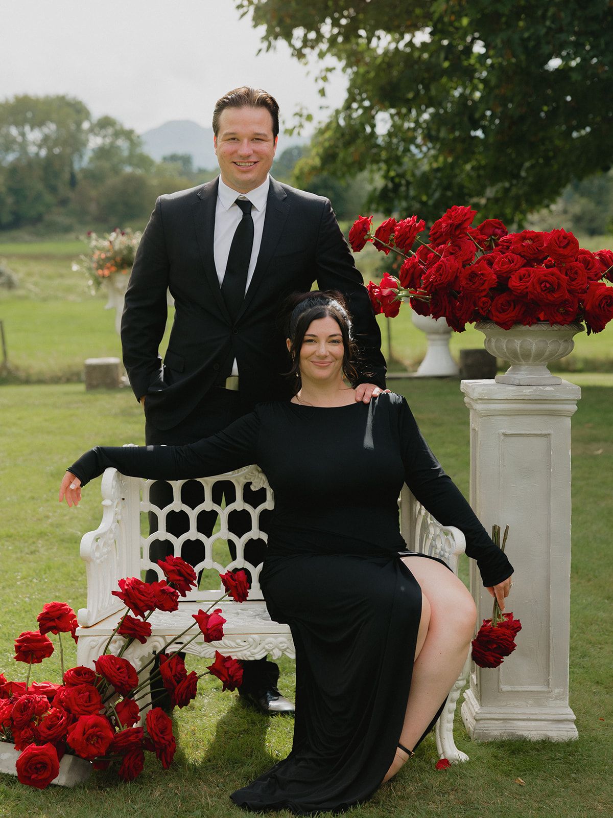 A couple sits on a white stone bench surrounded by vibrant red roses in an outdoor garden setting.