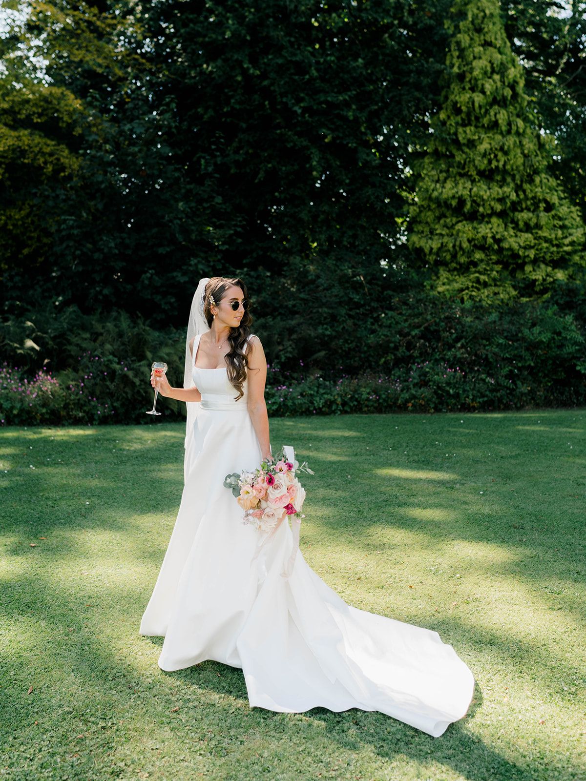 A bride in a flowing white wedding gown with long train stands on a sunlit lawn surrounded by lush green trees.