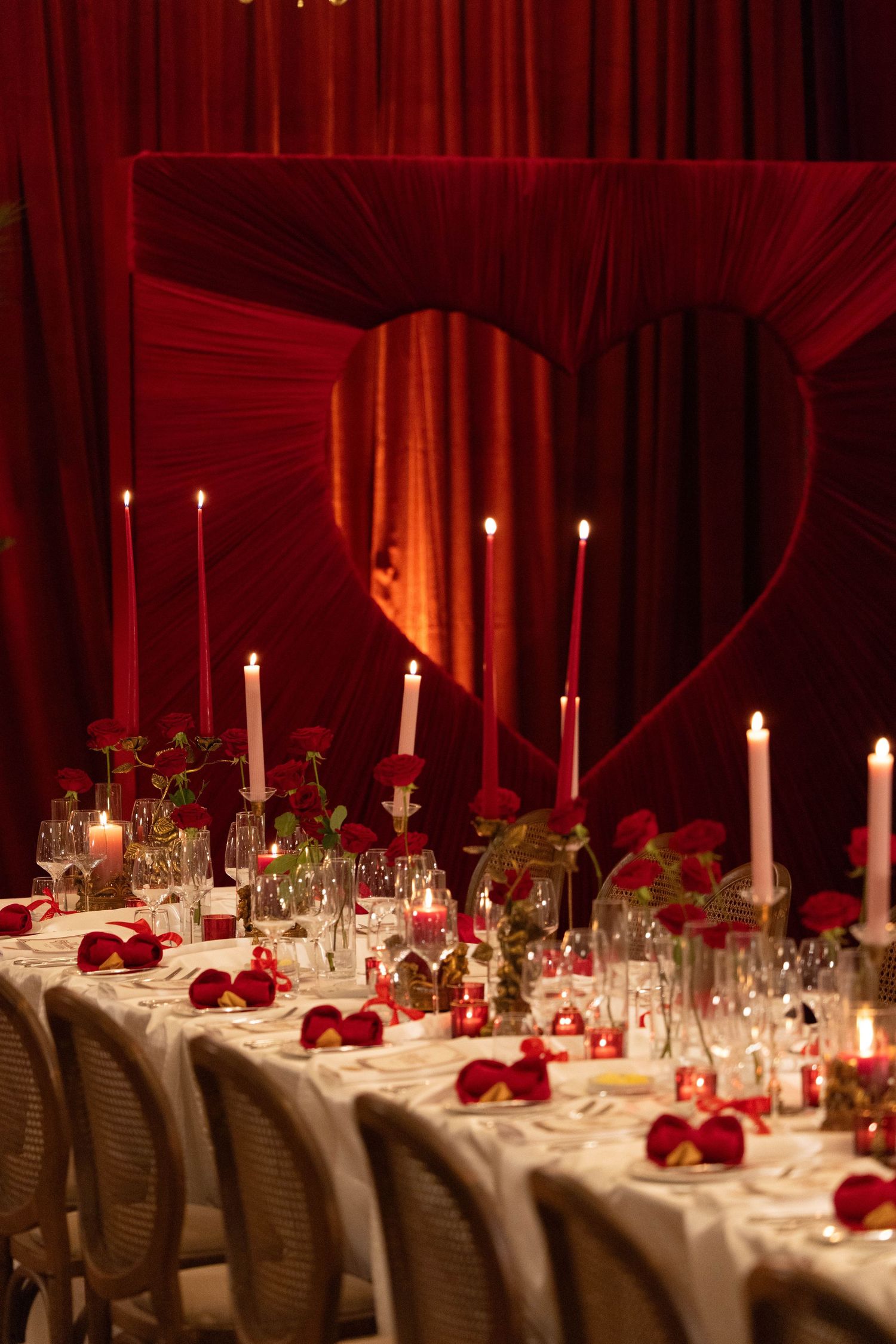 Elegant Valentine's Day dinner table with red roses, candles, and heart-shaped drape backdrop in romantic red lighting.