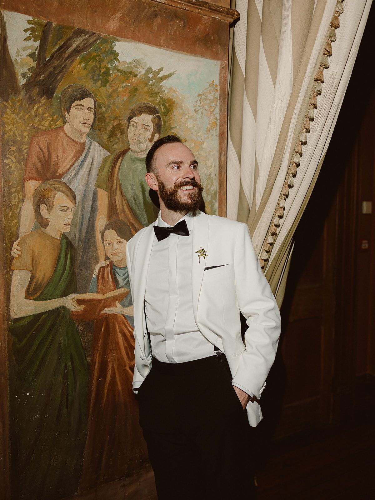 A person in a crisp white tuxedo jacket and bowtie smiles while standing next to a vintage wall mural.