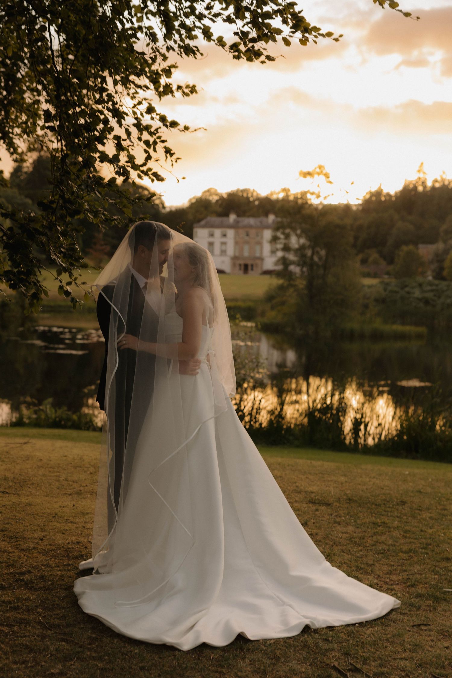 A couple in wedding attire embraces during sunset by a lake, creating romantic silhouettes against the golden sky.