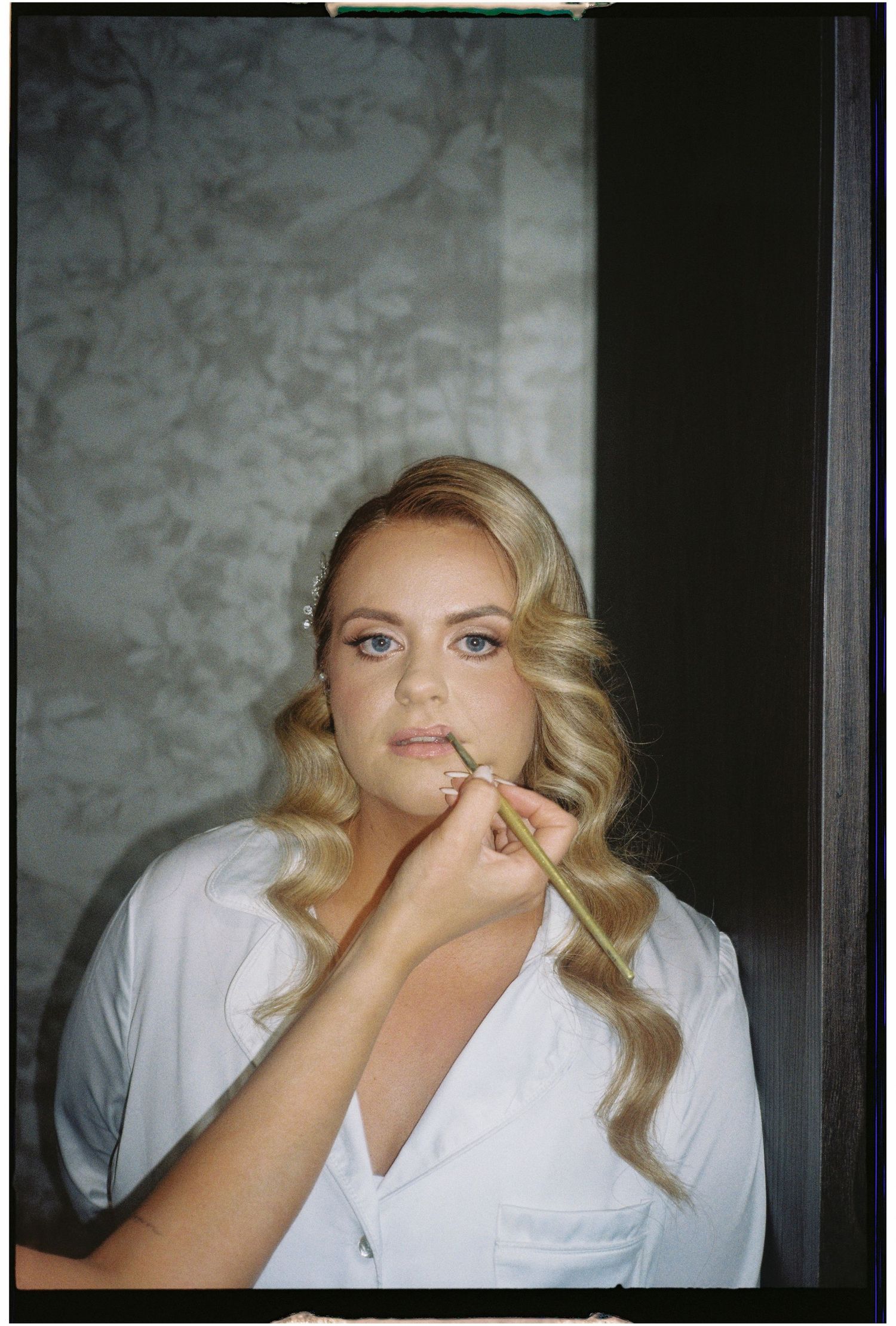 A person with blonde wavy hair wearing a white button-up shirt poses against a textured wall in moody lighting.