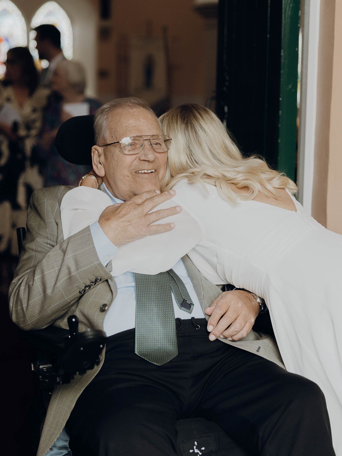 An emotional embrace between a senior man in a wheelchair wearing a gray suit and tie and a person in white.