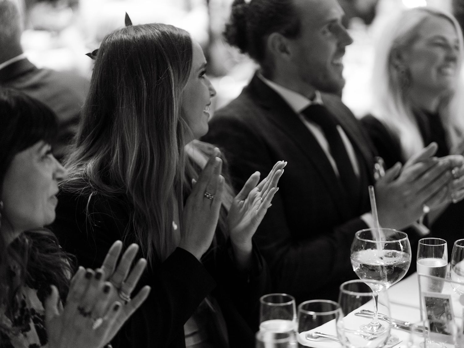A black and white photograph shows people clapping while seated at a formal dinner with wine glasses on the table.