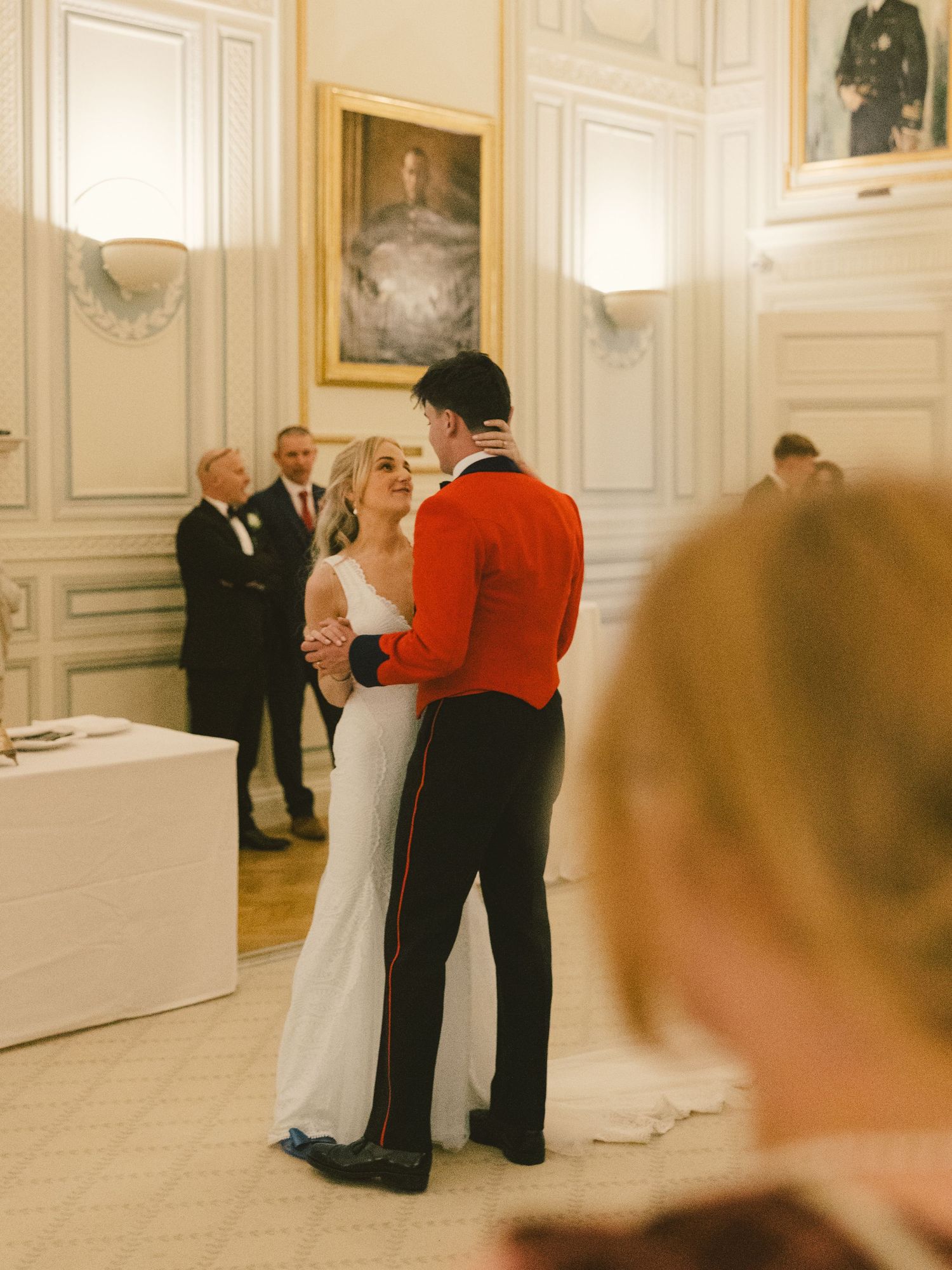 Guests in formal attire mingle in an elegant room with ornate white walls and trim during an upscale event.
